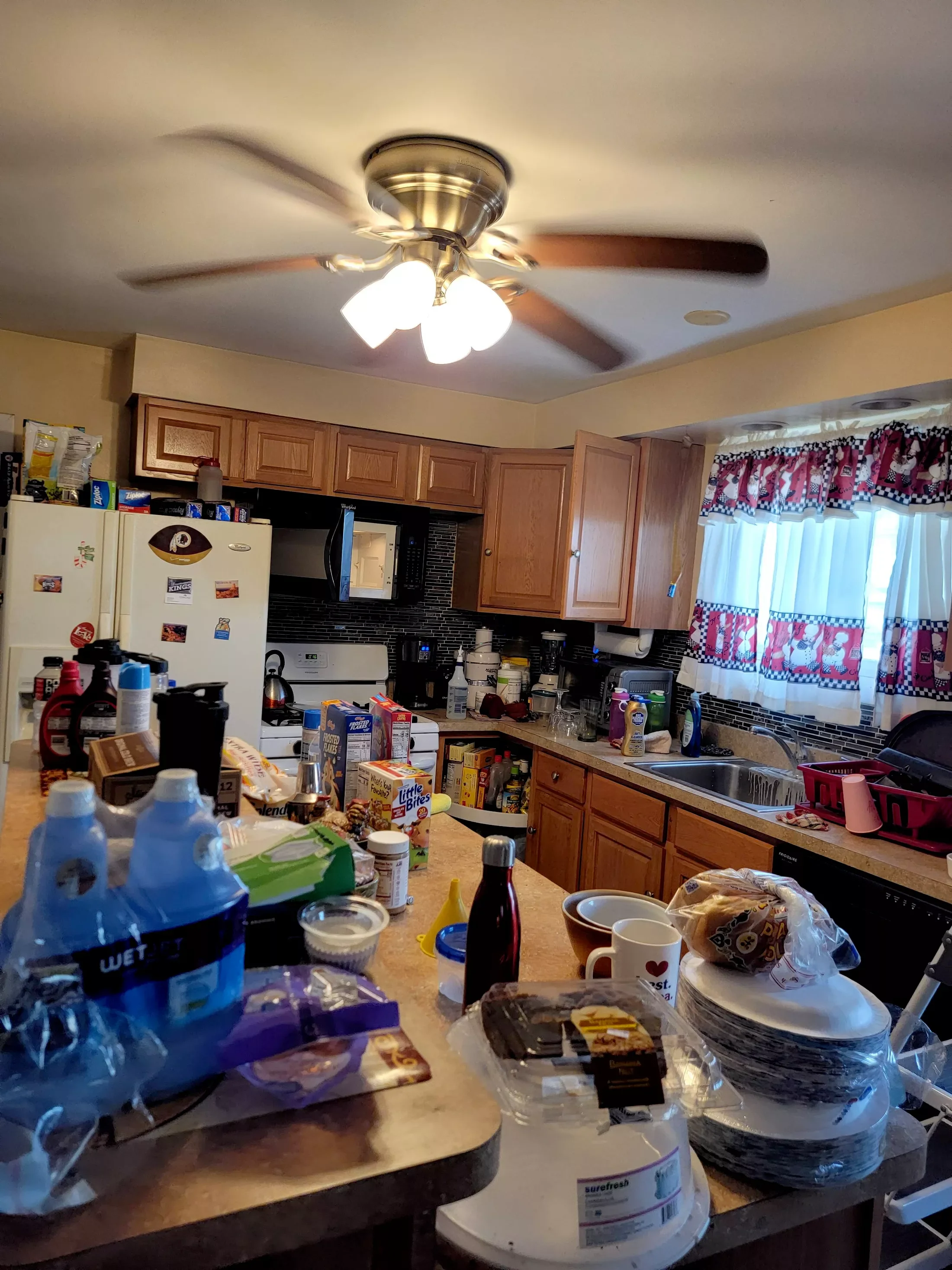 A cluttered kitchen with wooden cabinets, a countertops filled with various food items, drinks, containers, and utensils, a white refrigerator with magnets and items on top, a window with red and white curtains, and a ceiling fan with four lights.