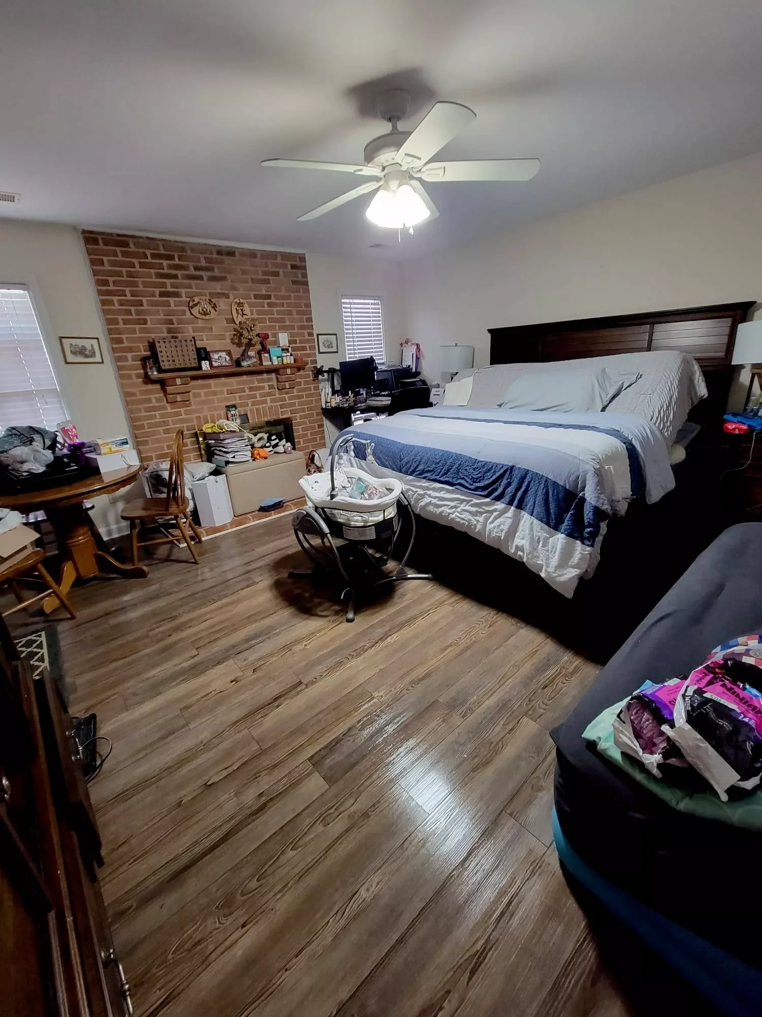 A cluttered bedroom with a large bed, brick fireplace, ceiling fan, wooden floor, and various furniture and household items.