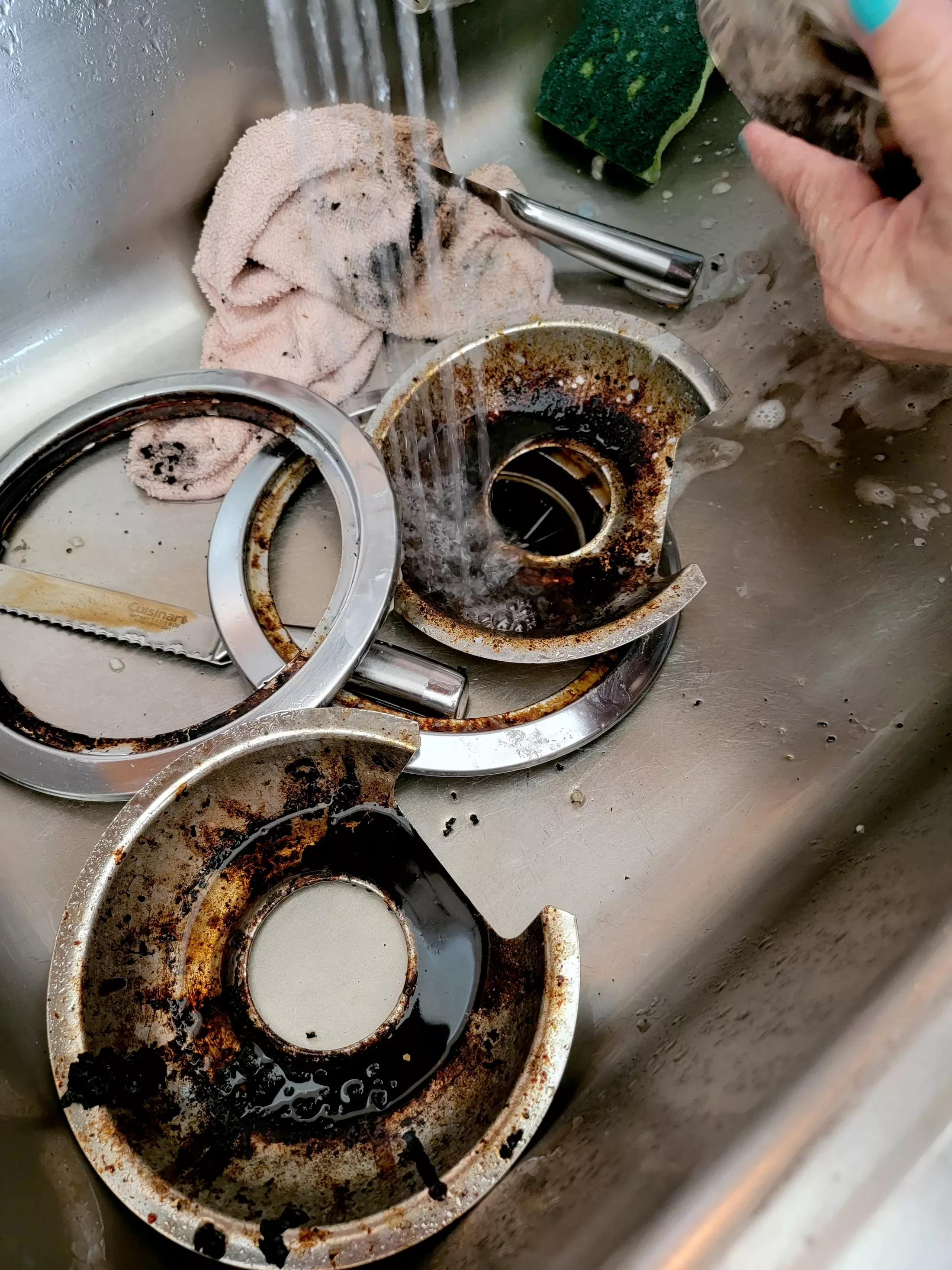 Dirty, greasy stovetop burners and parts in a kitchen sink, with a hand holding a scrub brush, soap, and a sponge nearby.