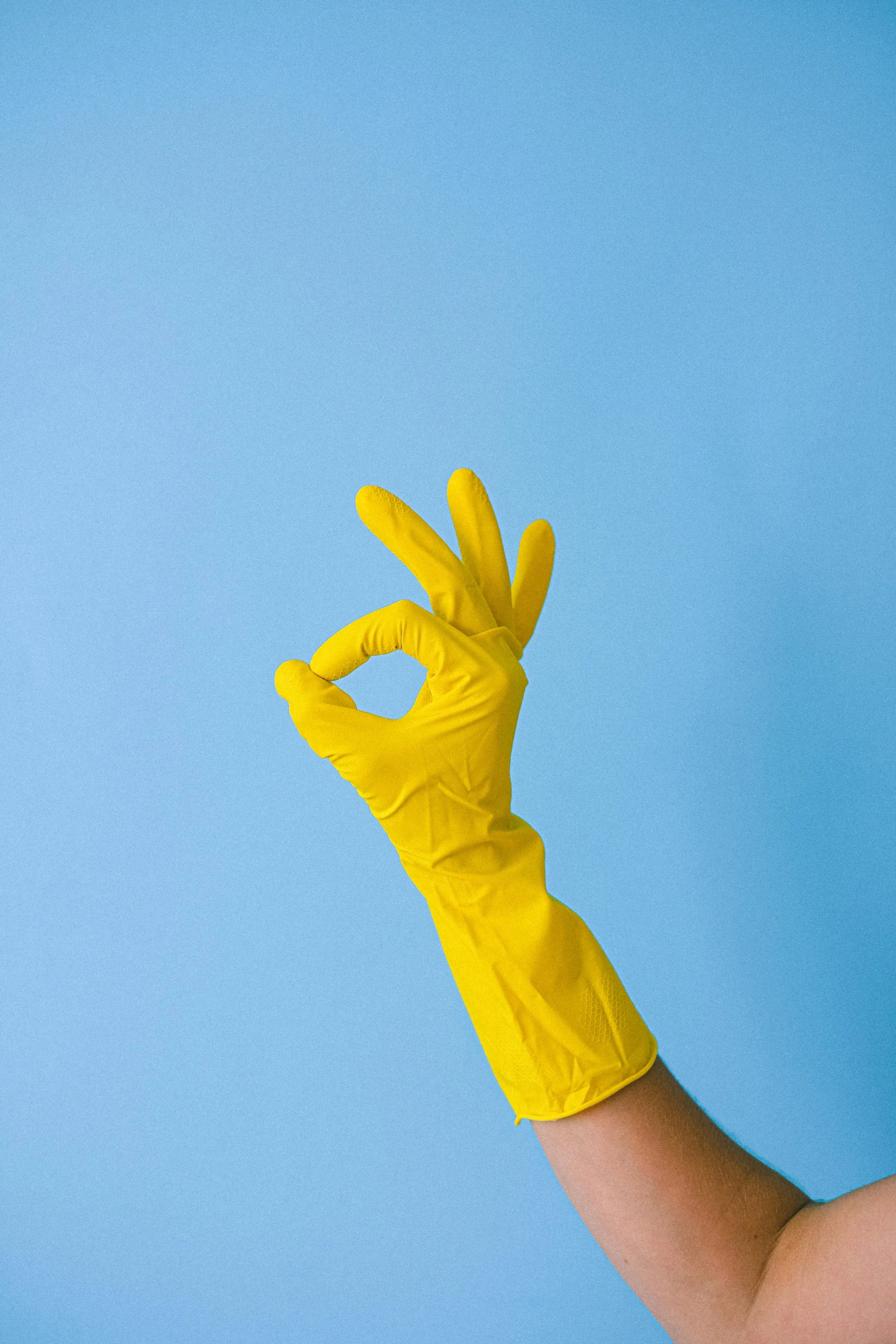 A hand wearing a yellow rubber glove making an okay sign against a light blue background.