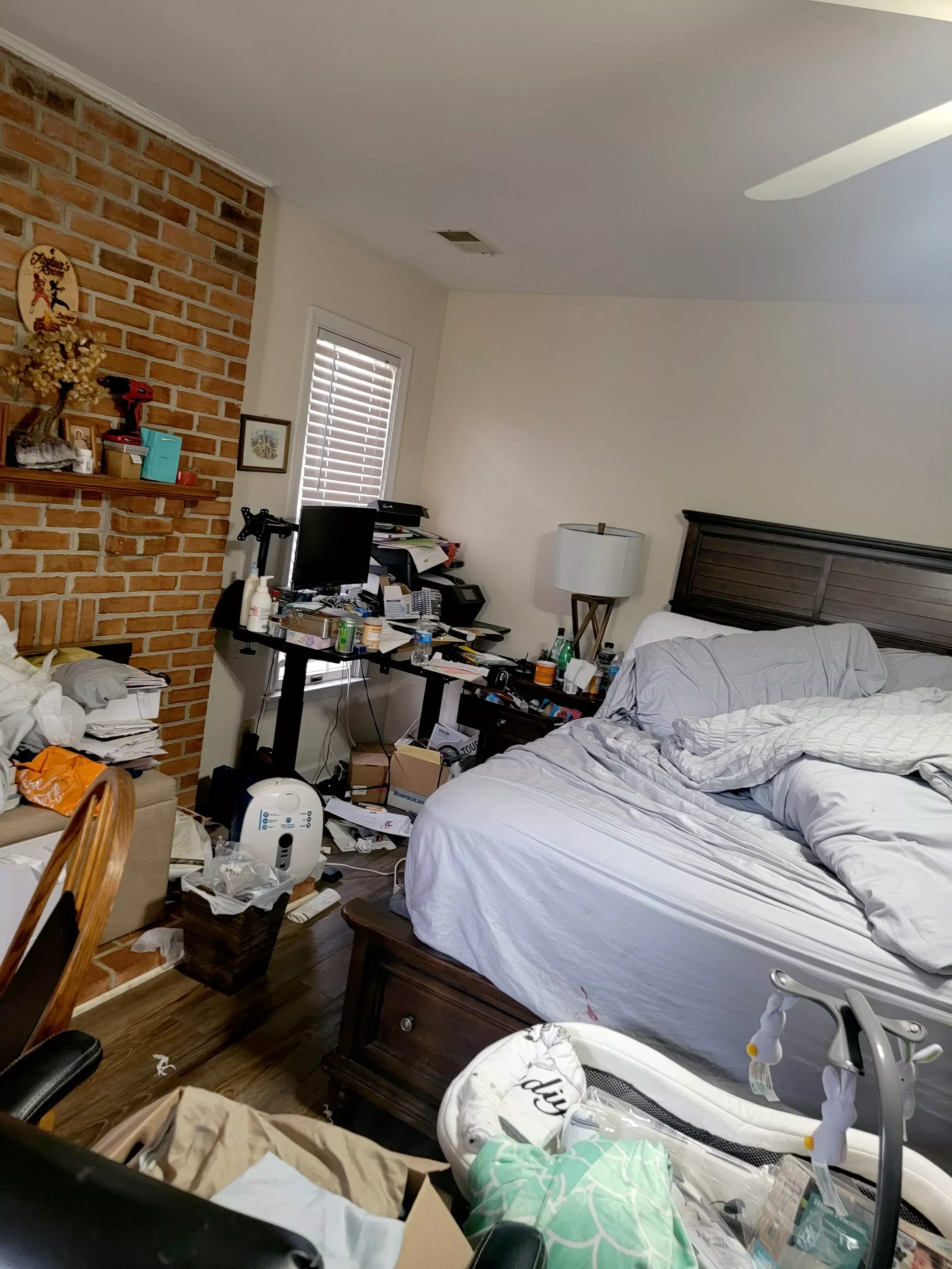A cluttered bedroom with an unmade bed, a messy desk with a computer and papers, a brick fireplace, and various items scattered around, including a lamp, bottles, and boxes.