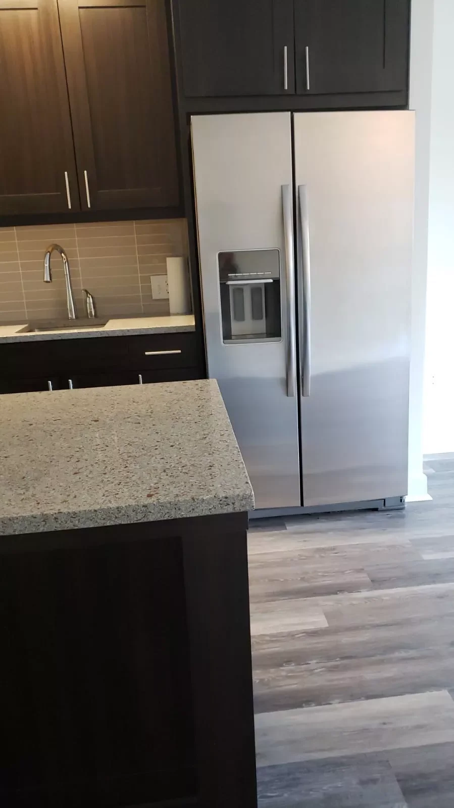 A modern kitchen featuring a stainless steel side-by-side refrigerator with a water and ice dispenser, dark wood cabinets, a light-colored granite countertop island, and a gray tile backsplash. The floor has a wood-like appearance.