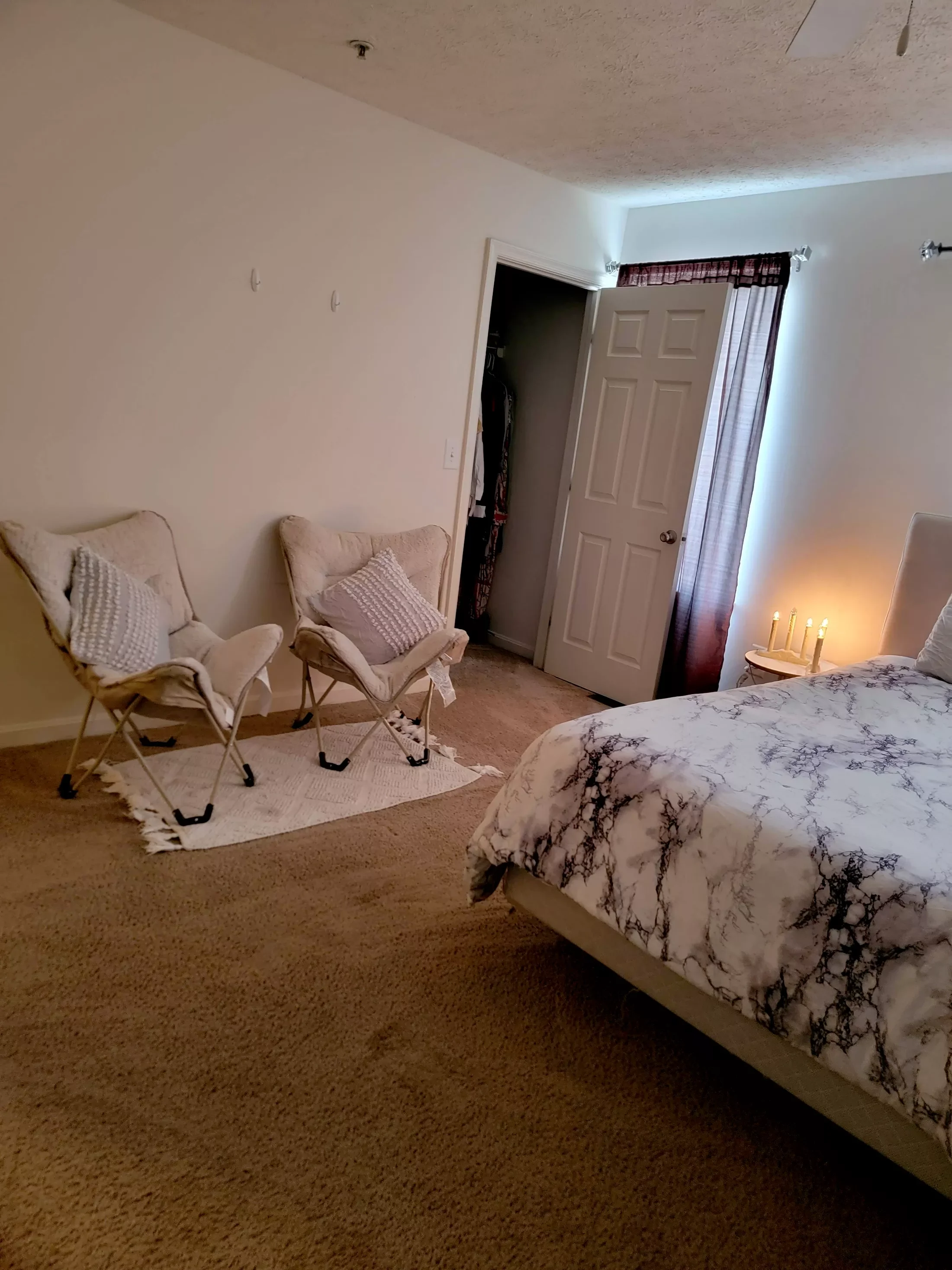 A bedroom with two cream-colored chairs, a bed with marble-patterned bedding, a small round nightstand with candles, and a door with a curtain.