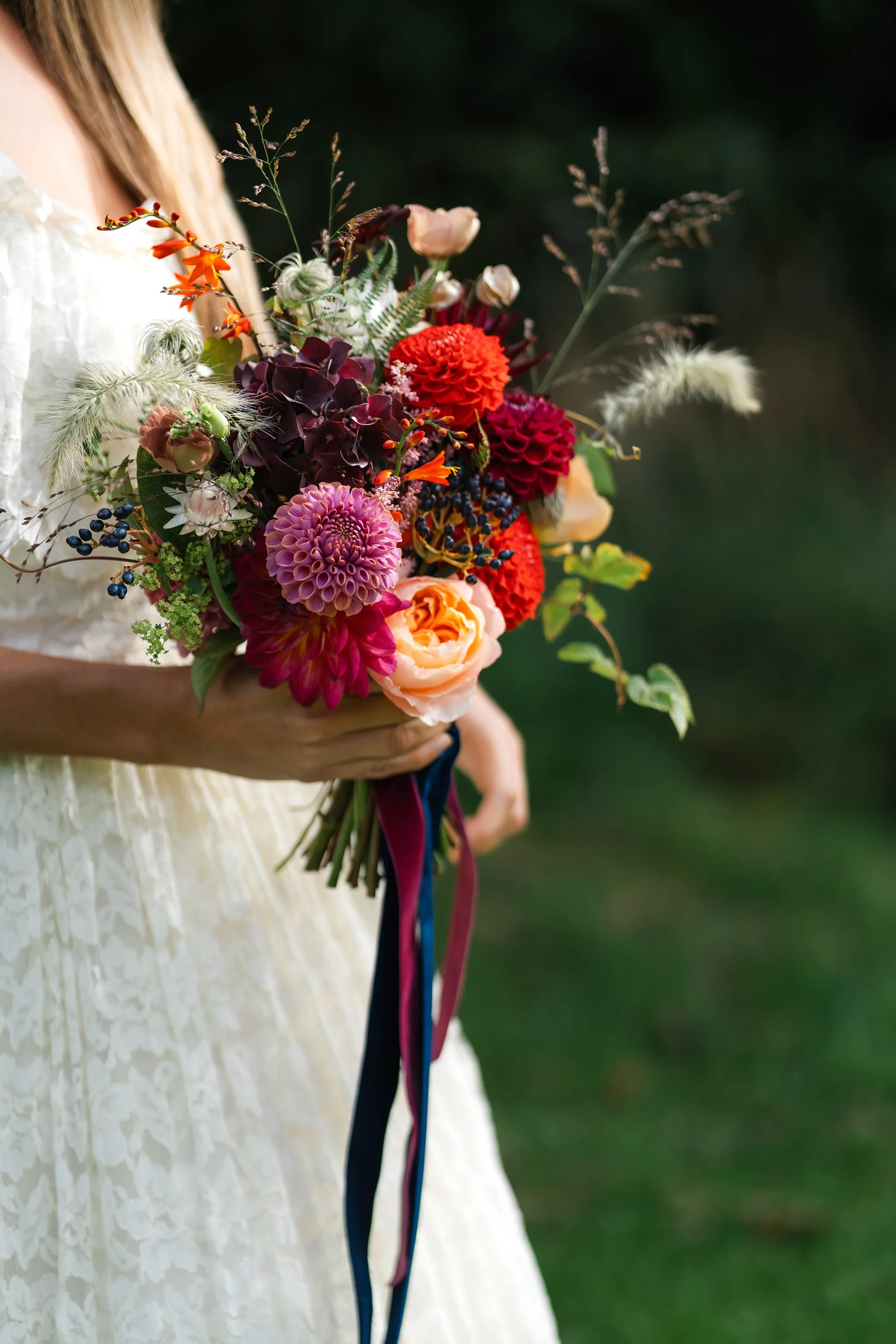 A person dressed in a cream-colored lace dress holding a bouquet of colorful flowers, including purple, red, pink, orange, and white blooms with greenery and ribbons, outdoors with a blurred natural green background.
