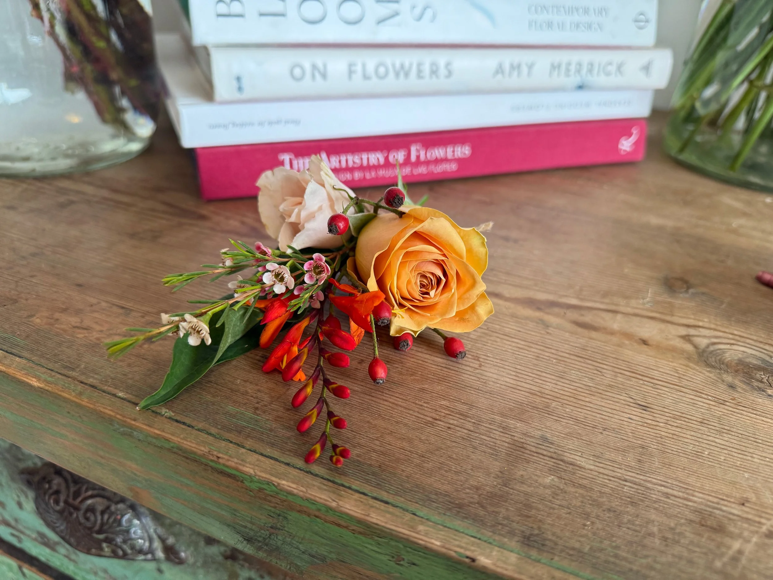 A small arrangement of flowers, including an orange rose, pink and white waxflower, red berries, and green foliage, placed on a rustic wooden surface in front of a stack of books.