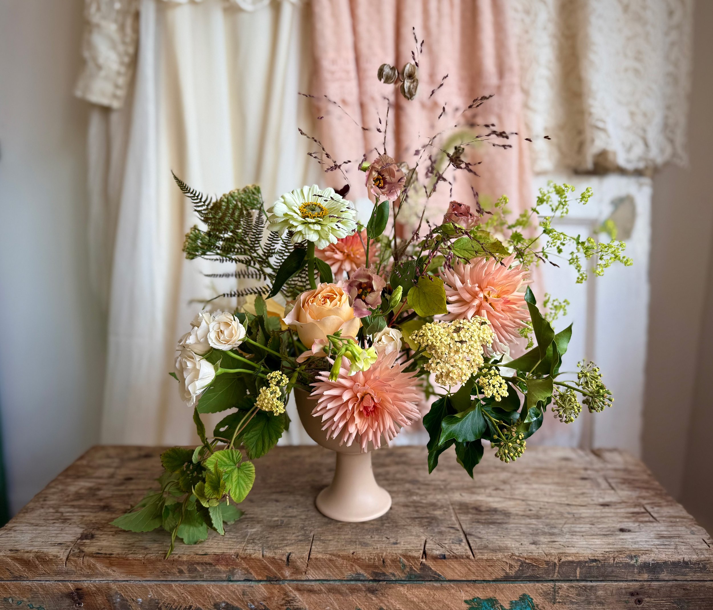 A floral arrangement in a white vase with pink, white, and peach flowers, green leaves, and sprigs of lavender, placed on a rustic wooden surface with a textured beige and pink background.