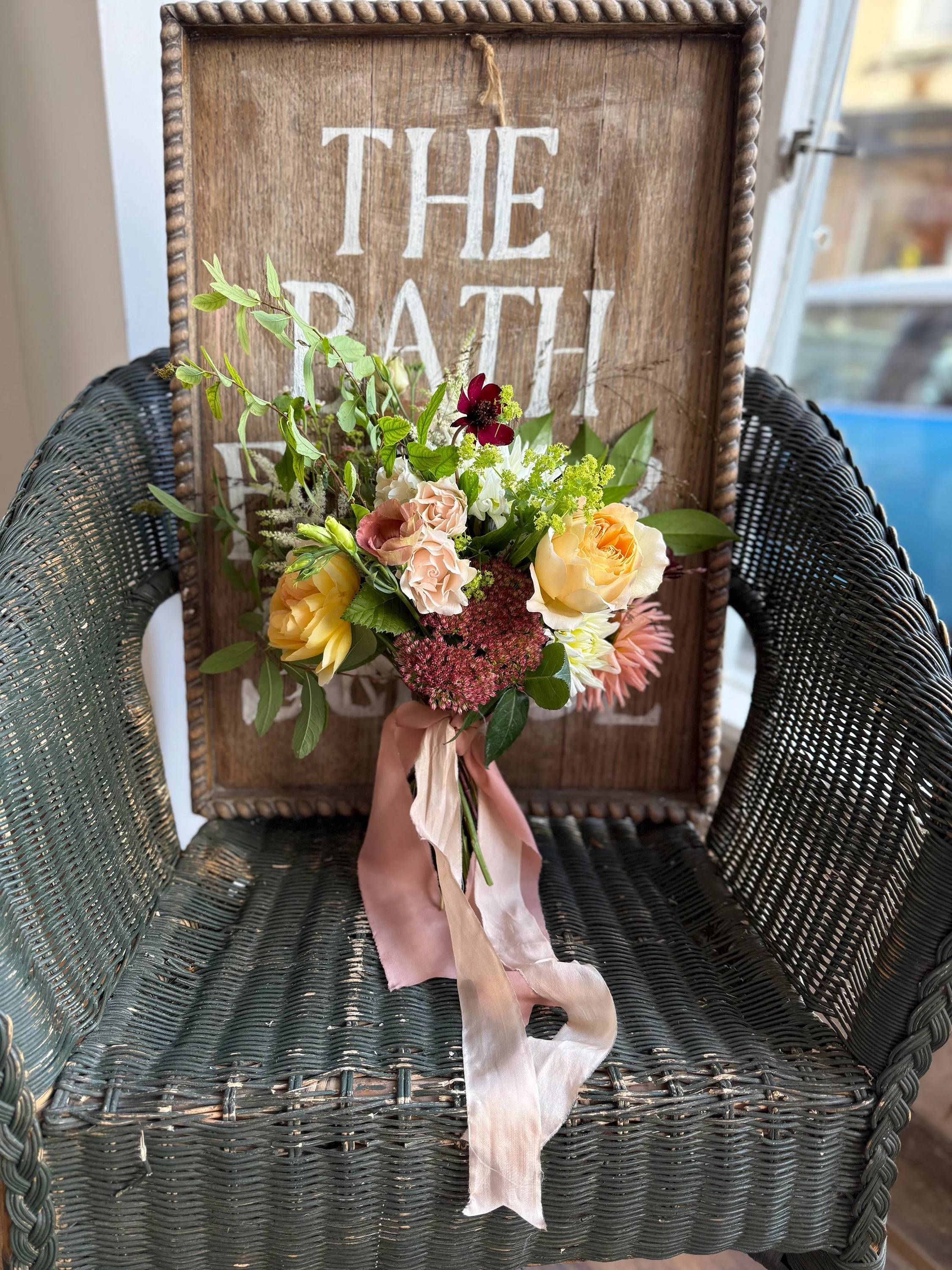 A bouquet of mixed flowers including roses and greenery is held in front of a wooden sign that reads 'The Bath' in white letters. The bouquet has a pink ribbon tied around the stems.