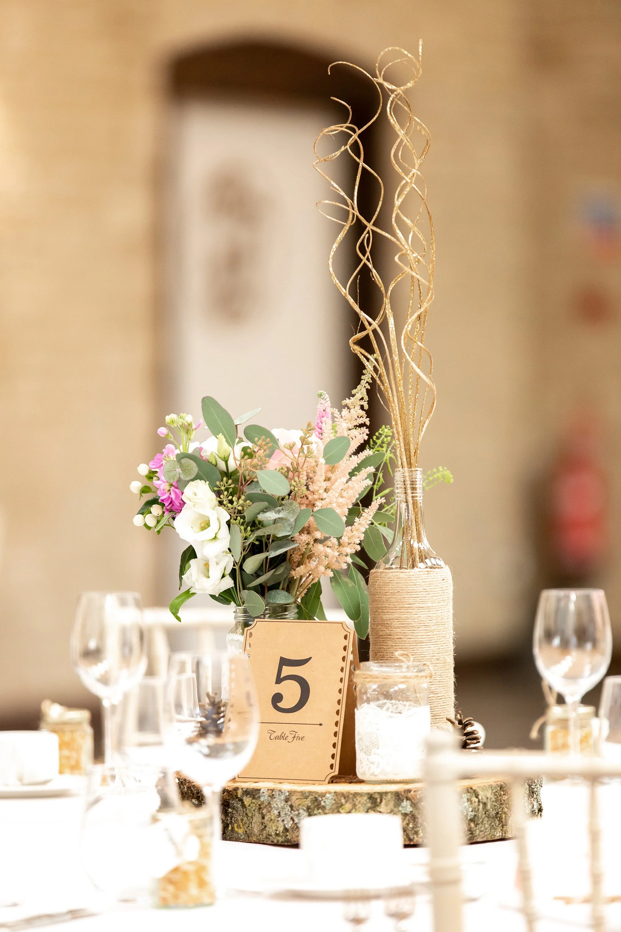Table centerpiece featuring a bouquet of flowers with greens, a gold glitter swirl decoration in a bottle wrapped with twine, a wooden slice base, and a table number card marked '5' at an indoor event with wine glasses and candles surrounding it.
