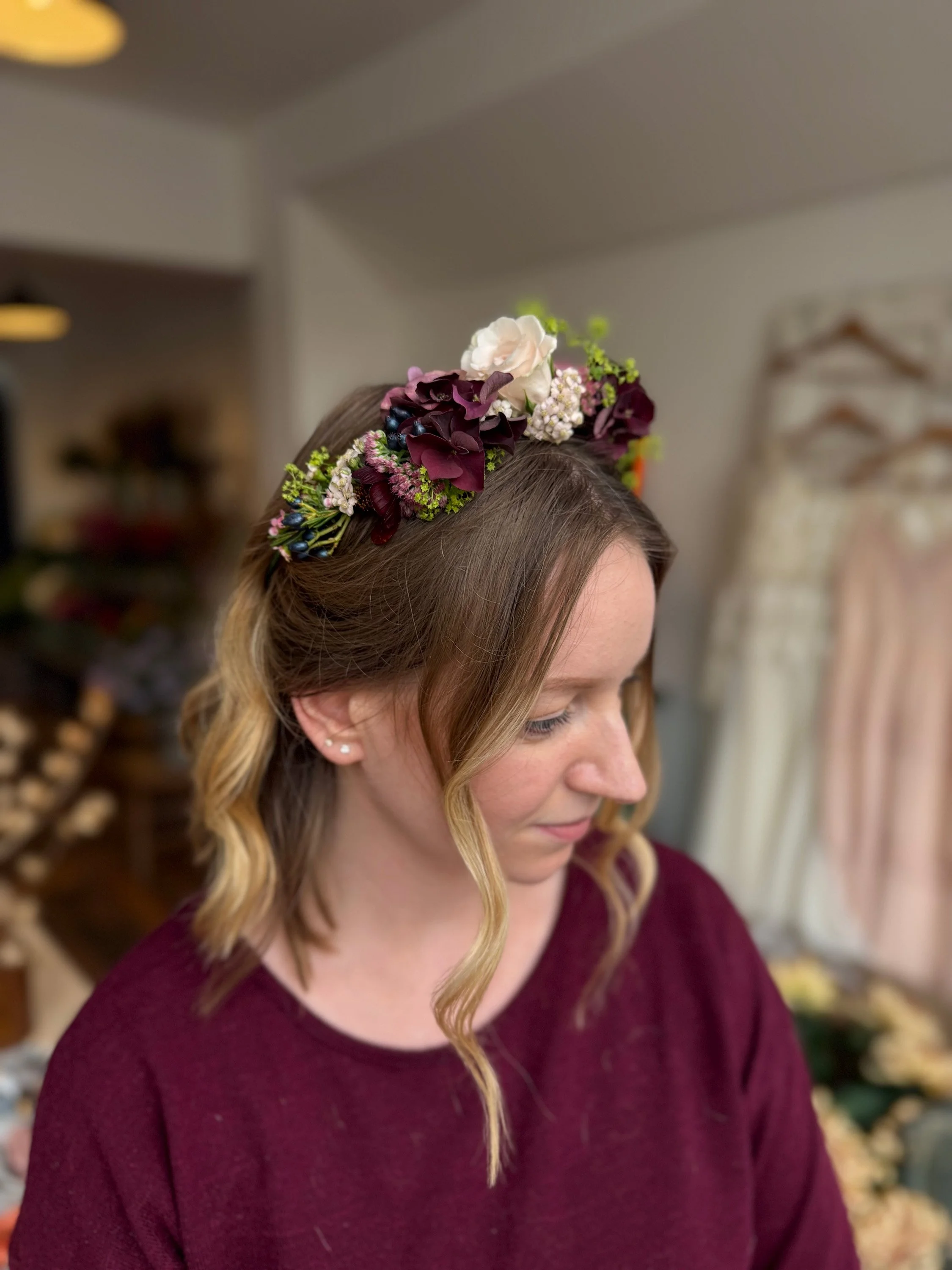 A woman with wavy, blonde hair wearing a burgundy sweater and a floral crown with white, pink, and purple flowers.