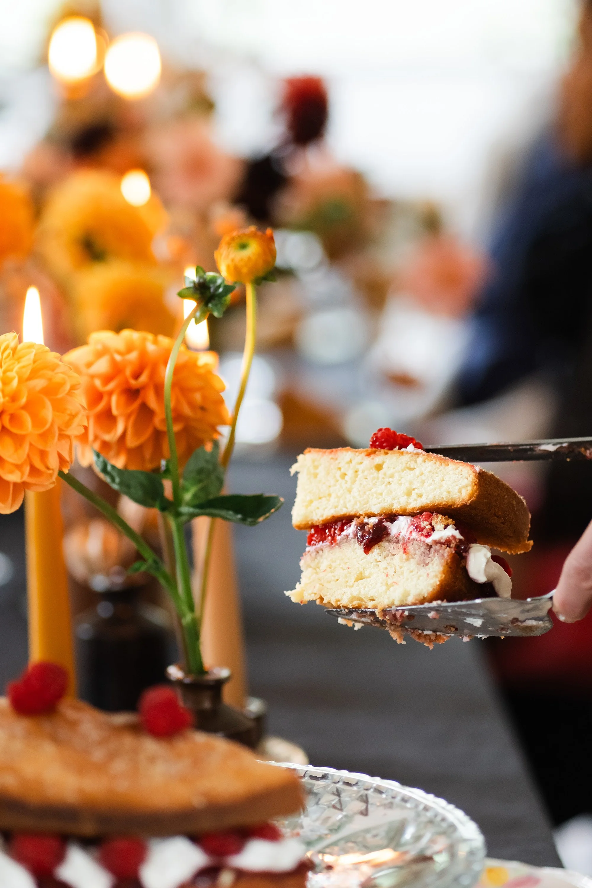 Close-up of a piece of cake on a spatula, with blurred colorful flowers and candles in the background at a celebration or party.
