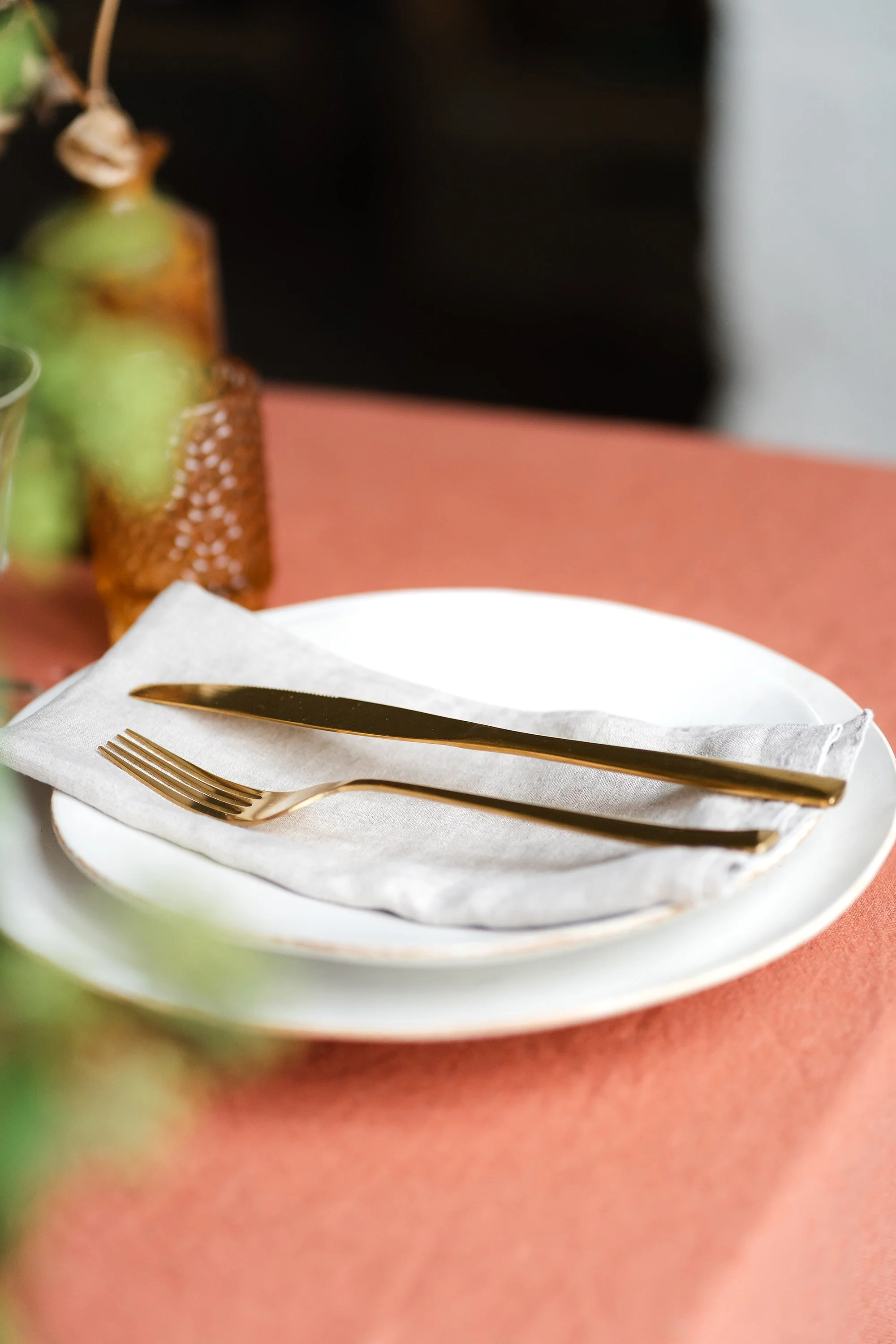 A white plate with a white napkin, gold-colored fork and knife on top, set on a pink table. In the background, a blurred image of a container with green grapes and other items.