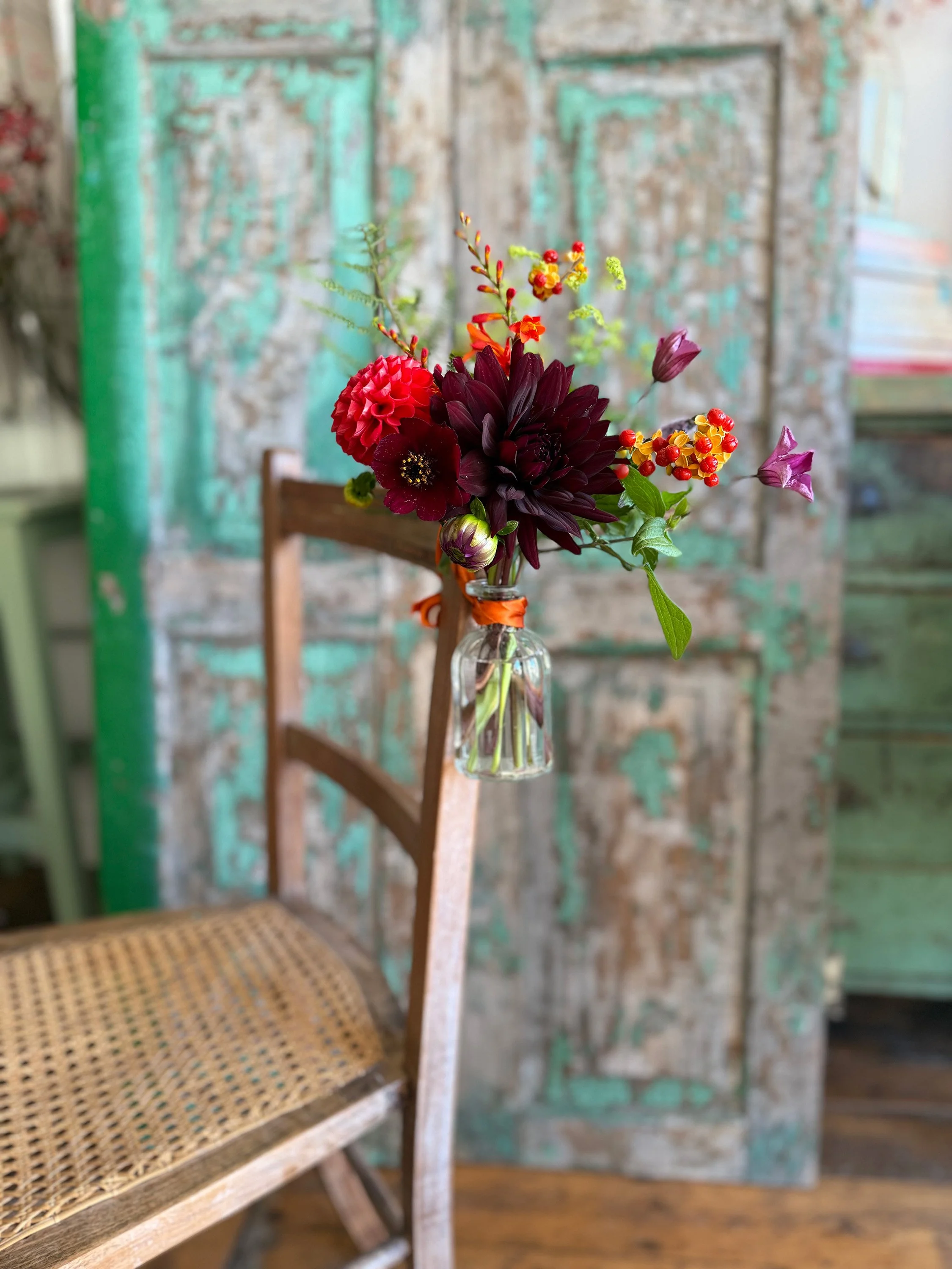 A glass vase with a colorful bouquet of flowers including dark purple, pink, red, orange, and yellow blooms, placed on a wooden chair with a woven seat, in front of a rustic textured wall with peeling paint.