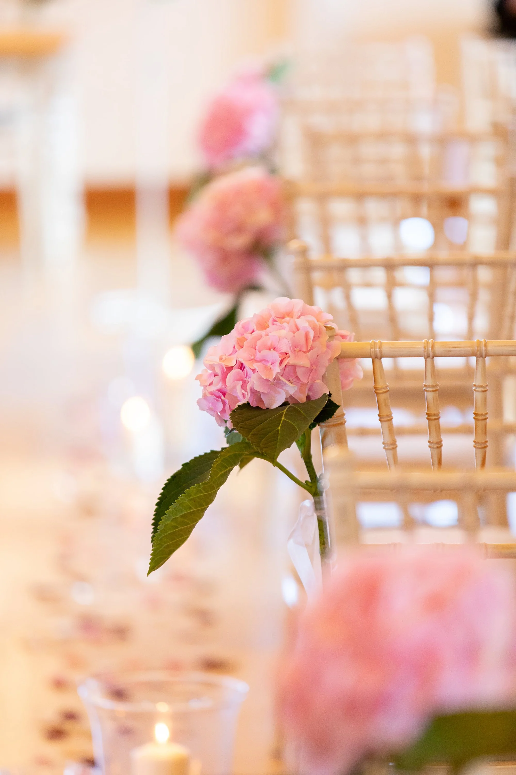 Close-up of a pink hydrangea flower with green leaves, attached to a chair at a decorated table setting with blurred background including candles.