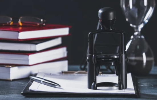Desk with a stack of red, white, and black books, a pair of glasses, a pen, a stamp, and an hourglass.