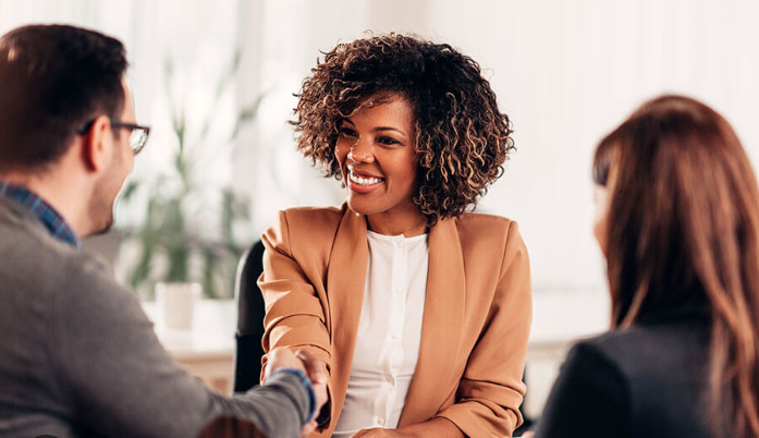 A woman with curly hair shaking hands with a man while another woman looks on in a professional setting.
