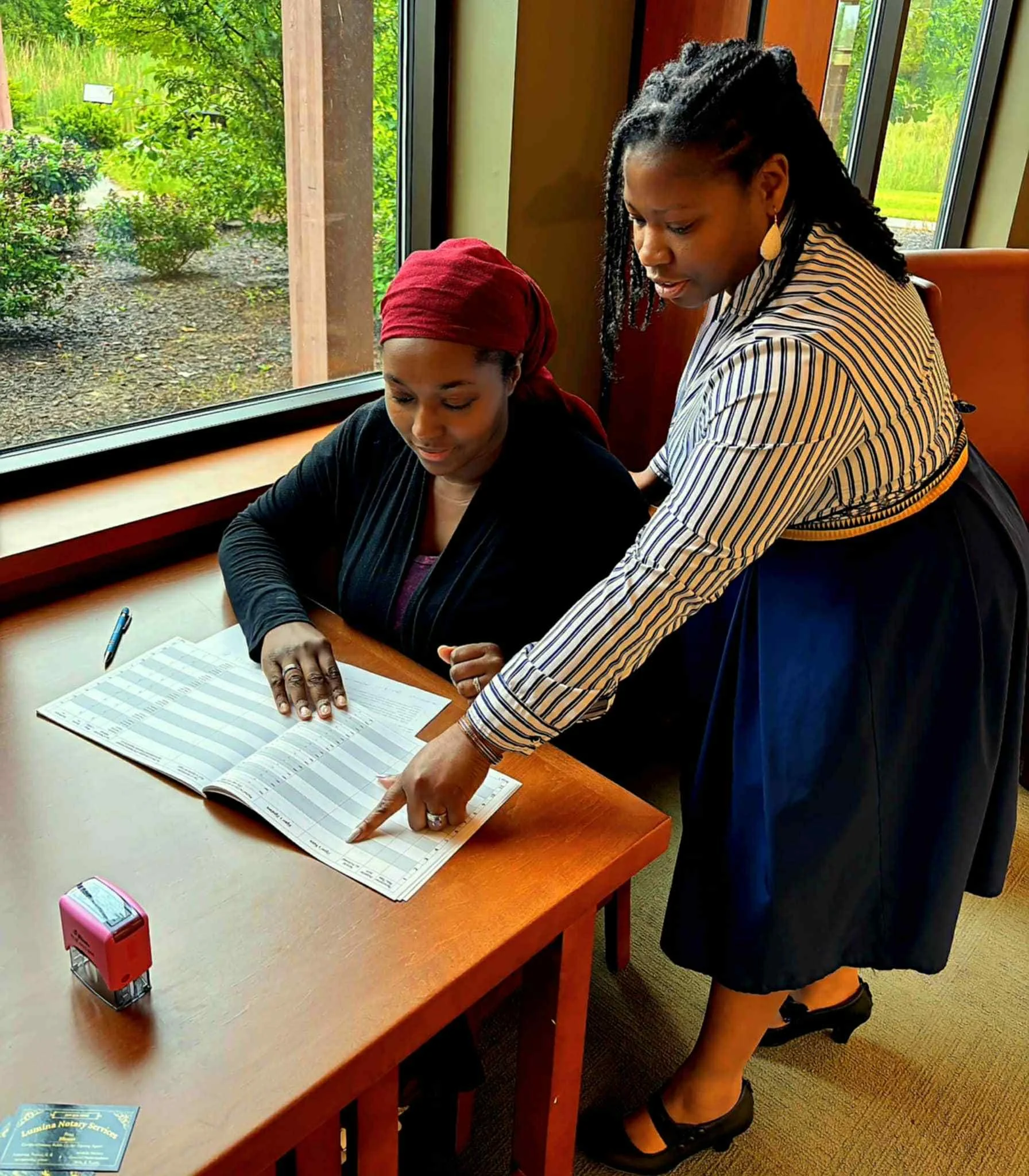 Two women in professional attire discussing a document at a wooden table near a window with a view of greenery outside.