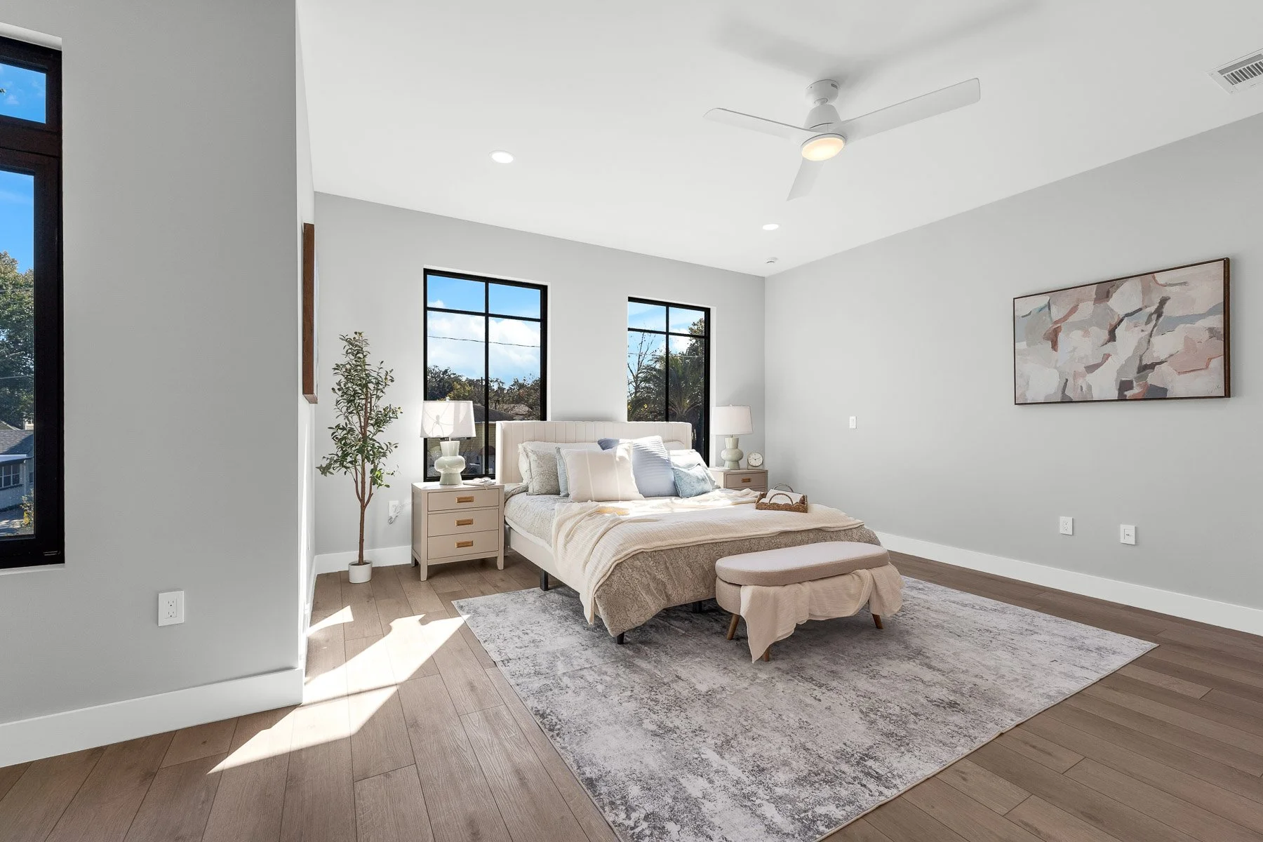 Modern living room with large windows, white walls, hardwood floors, a white armchair with a cushion, a small table, a floor lamp, and framed artwork. There is a doorway leading to another room, a plant, and a round white rug.
