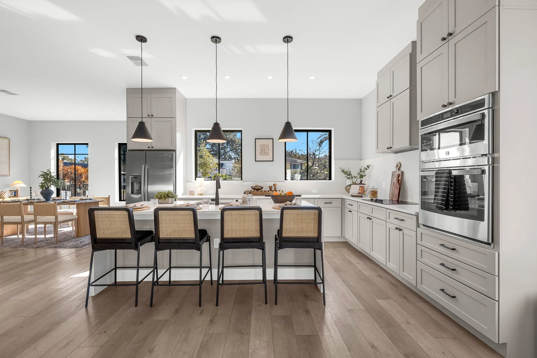 Modern home office with a wooden desk, two rattan chairs, large window, abstract art on wall, potted plant, and a patterned rug on hardwood floor.