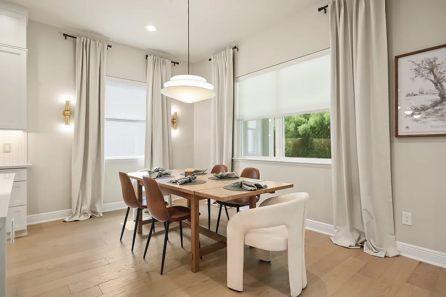 Modern living room with large windows, white walls, hardwood floors, a white armchair with a cushion, a small table, a floor lamp, and framed artwork. There is a doorway leading to another room, a plant, and a round white rug.