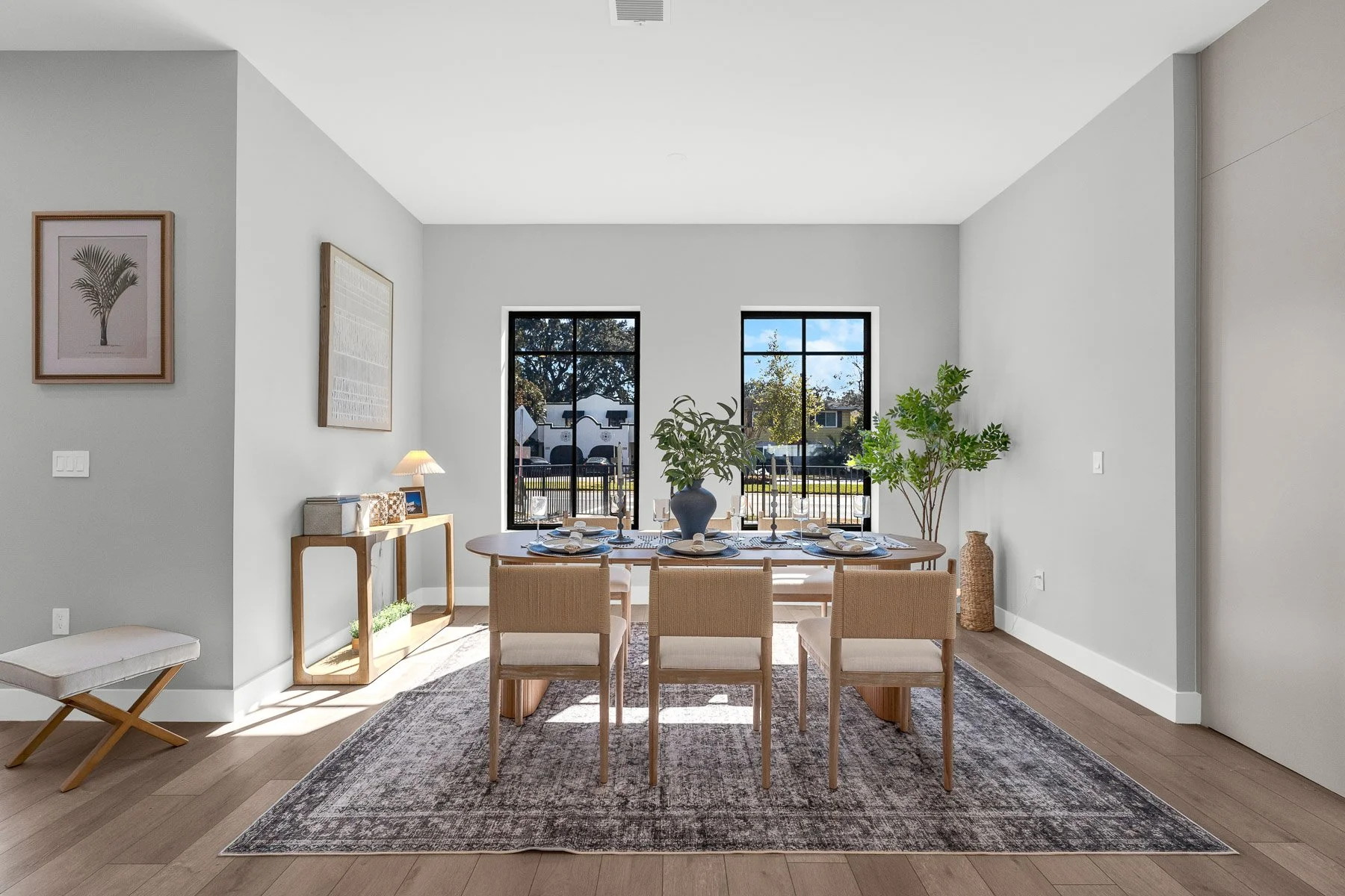 Bright bedroom with four large black-framed windows, light gray walls, a bed with gray bedding and throw pillows, nightstands with lamps, a wooden dresser, and a small bench near the window.