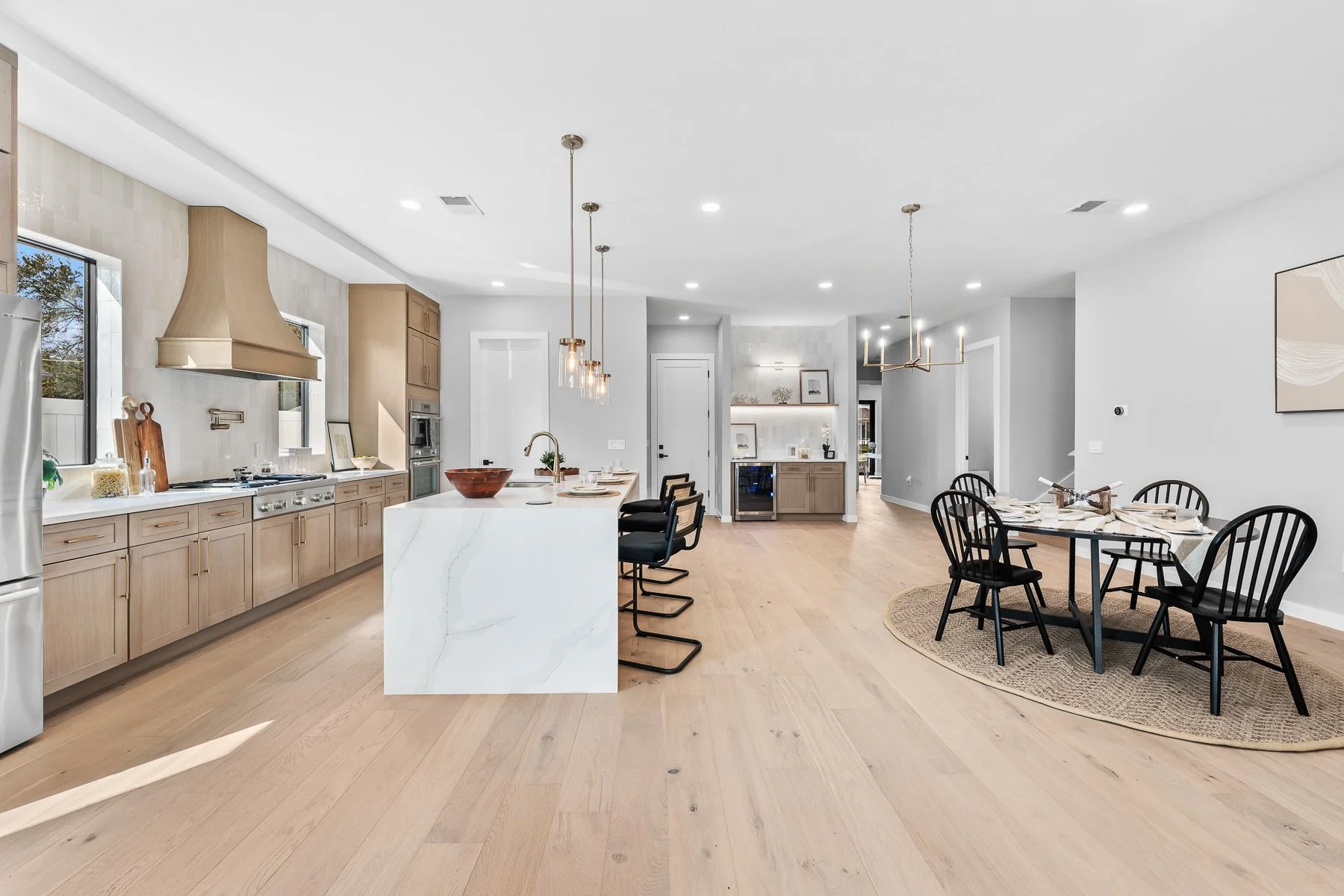 Modern living room with large windows, white walls, hardwood floors, a white armchair with a cushion, a small table, a floor lamp, and framed artwork. There is a doorway leading to another room, a plant, and a round white rug.