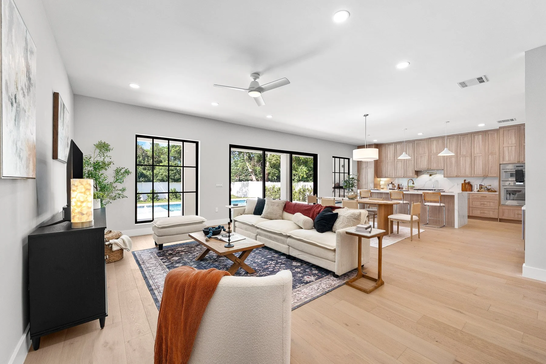 Modern living room with large windows, white walls, hardwood floors, a white armchair with a cushion, a small table, a floor lamp, and framed artwork. There is a doorway leading to another room, a plant, and a round white rug.