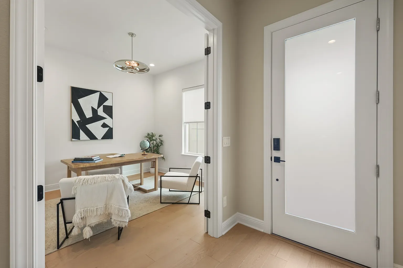 Modern home office with a wooden desk, two rattan chairs, large window, abstract art on wall, potted plant, and a patterned rug on hardwood floor.