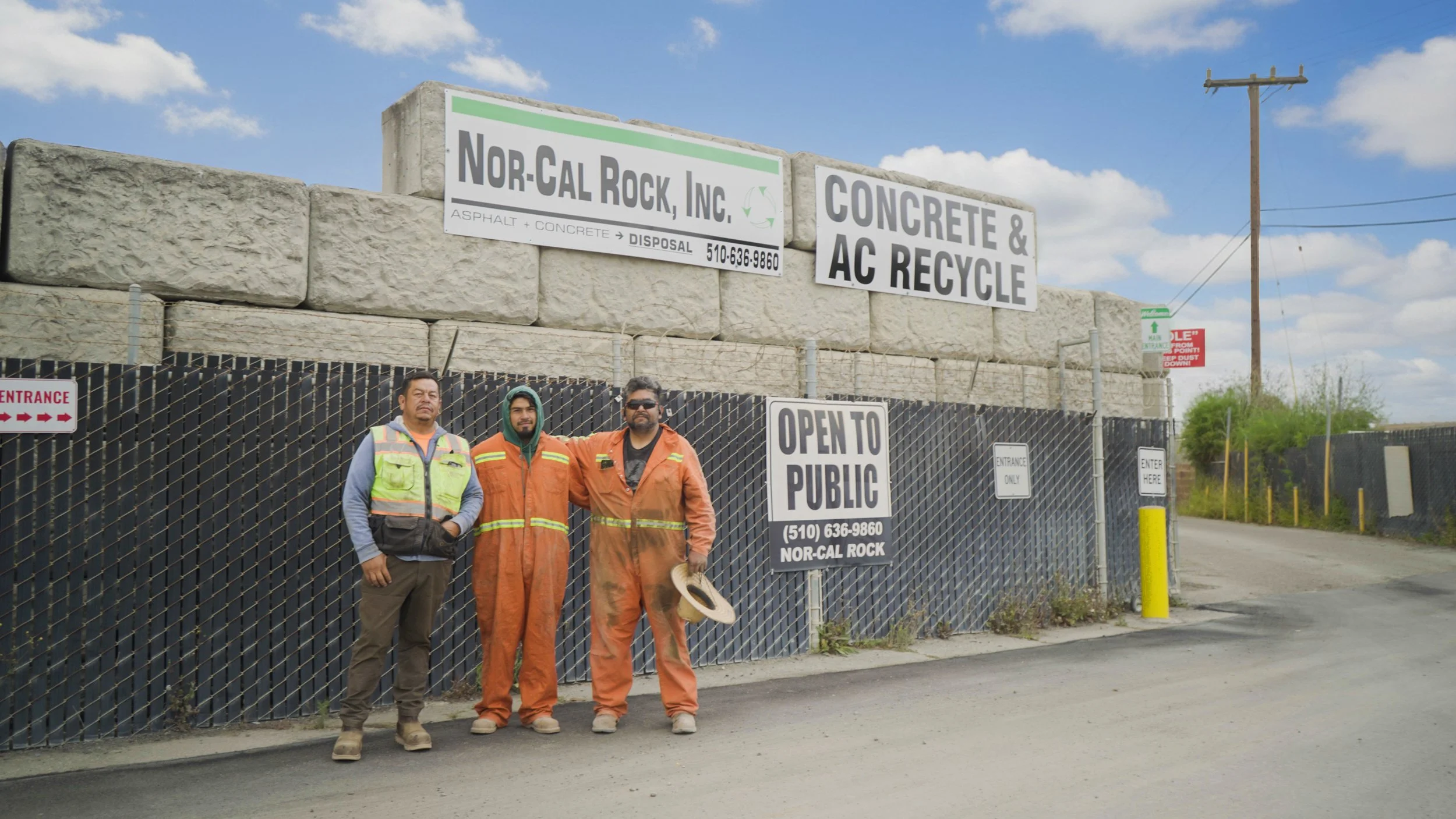 Three workers standing in front of a chain-link fence, with signs indicating it is a concrete and recycling business. The workers are wearing safety gear and standing on a dirt road under a partly cloudy sky.