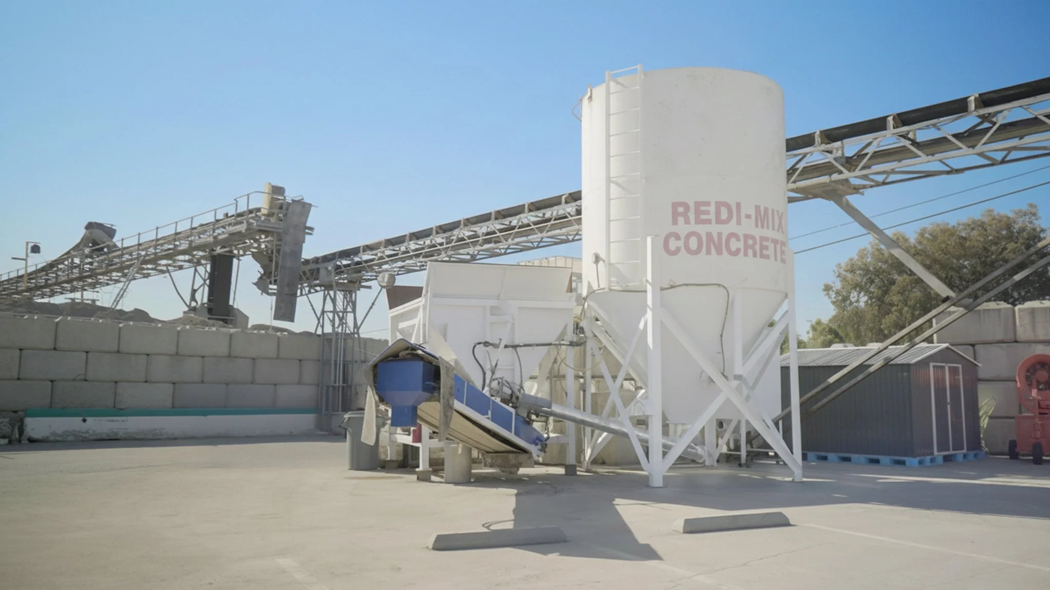Concrete mixing equipment, including a large white silo labeled 'REDI-MIX CONCRETE,' a conveyor belt, and a cement truck in the background, set against a blue sky and trees.