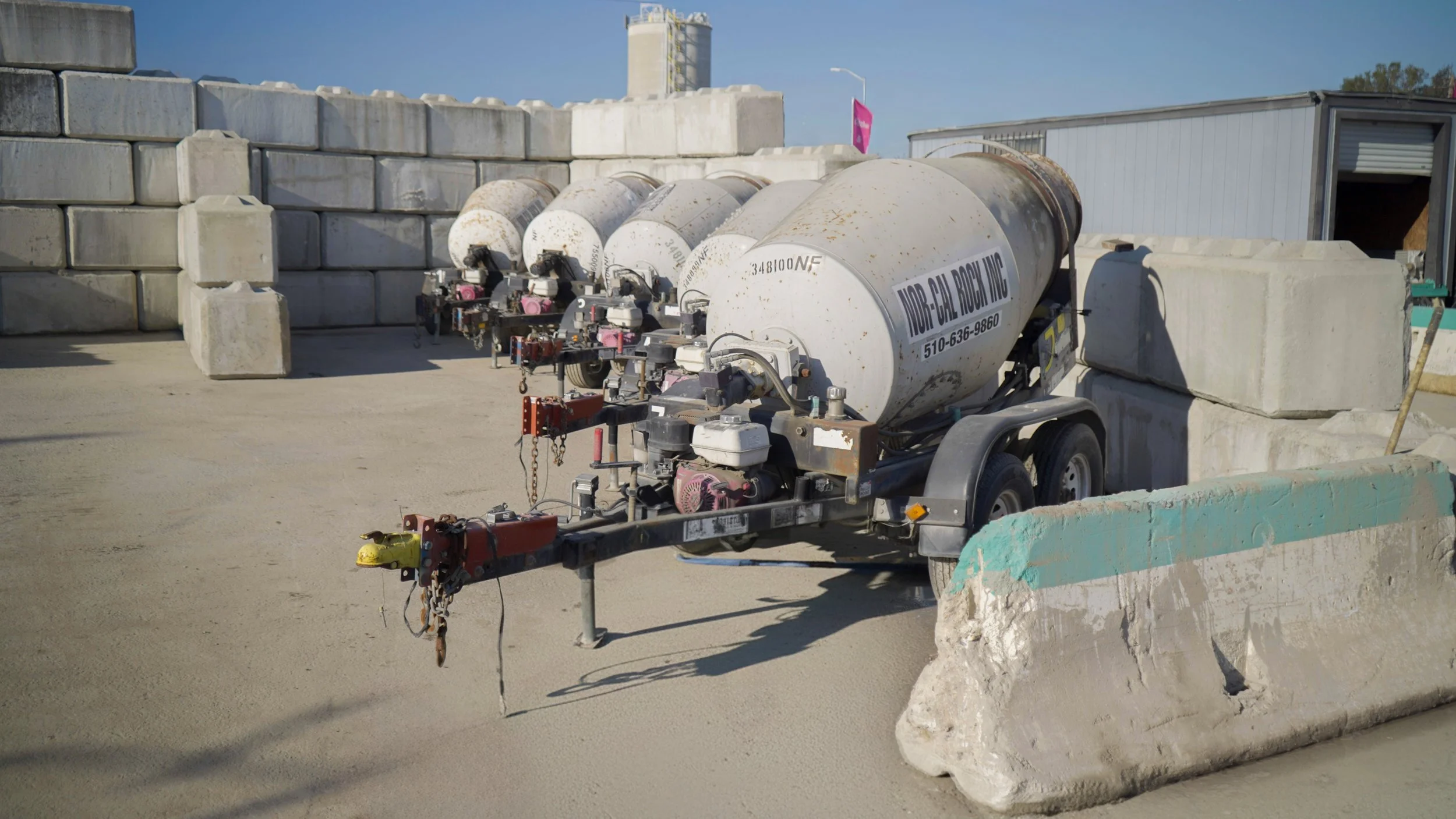 Concrete construction site with a trailer holding multiple large cement mixers, surrounded by concrete blocks and barriers.