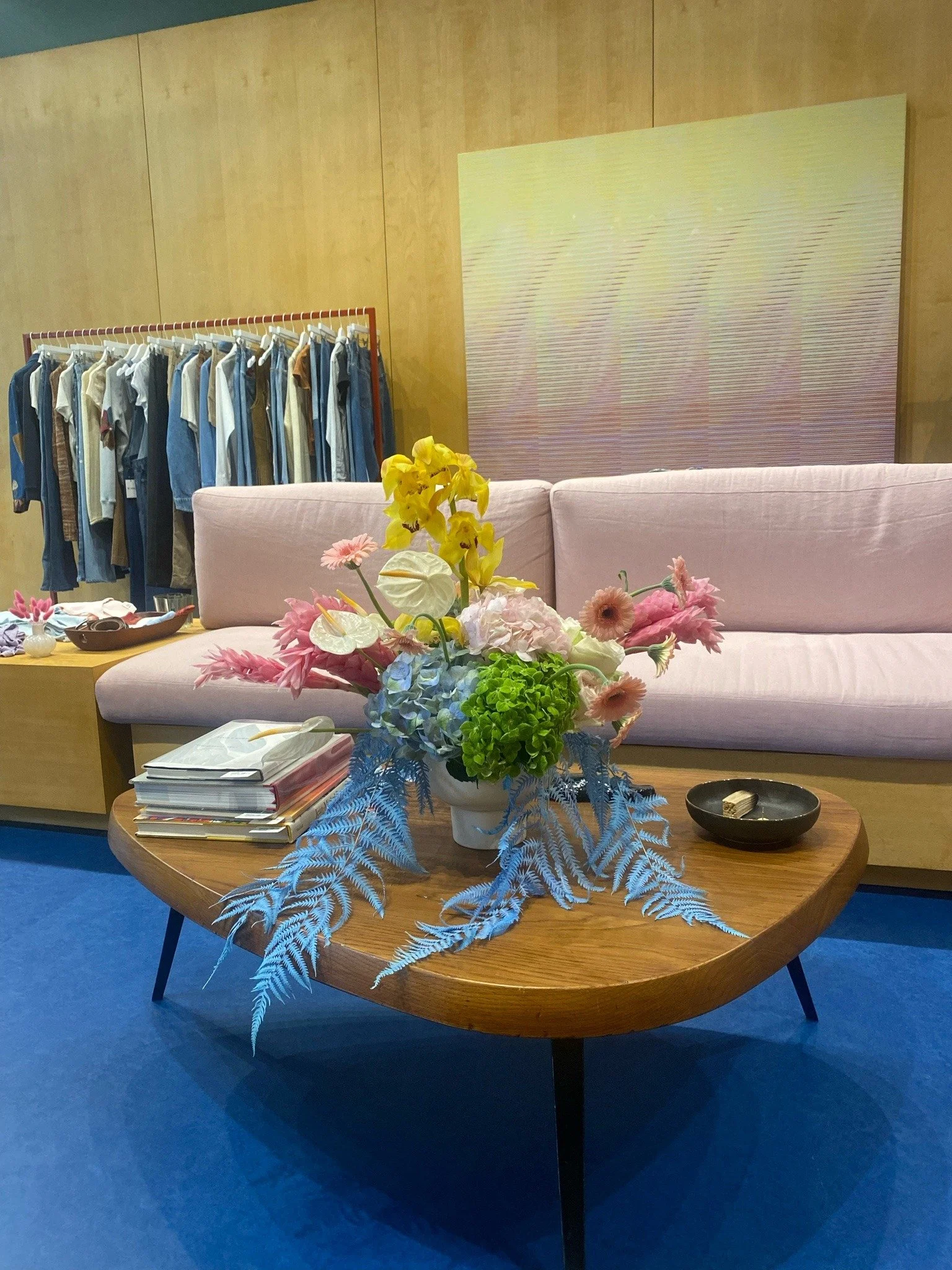 Living room setup with a pink sofa, a wooden coffee table with a colorful flower arrangement, a black bowl, and stacks of magazines, with a clothing rack filled with jeans and shirts in the background.