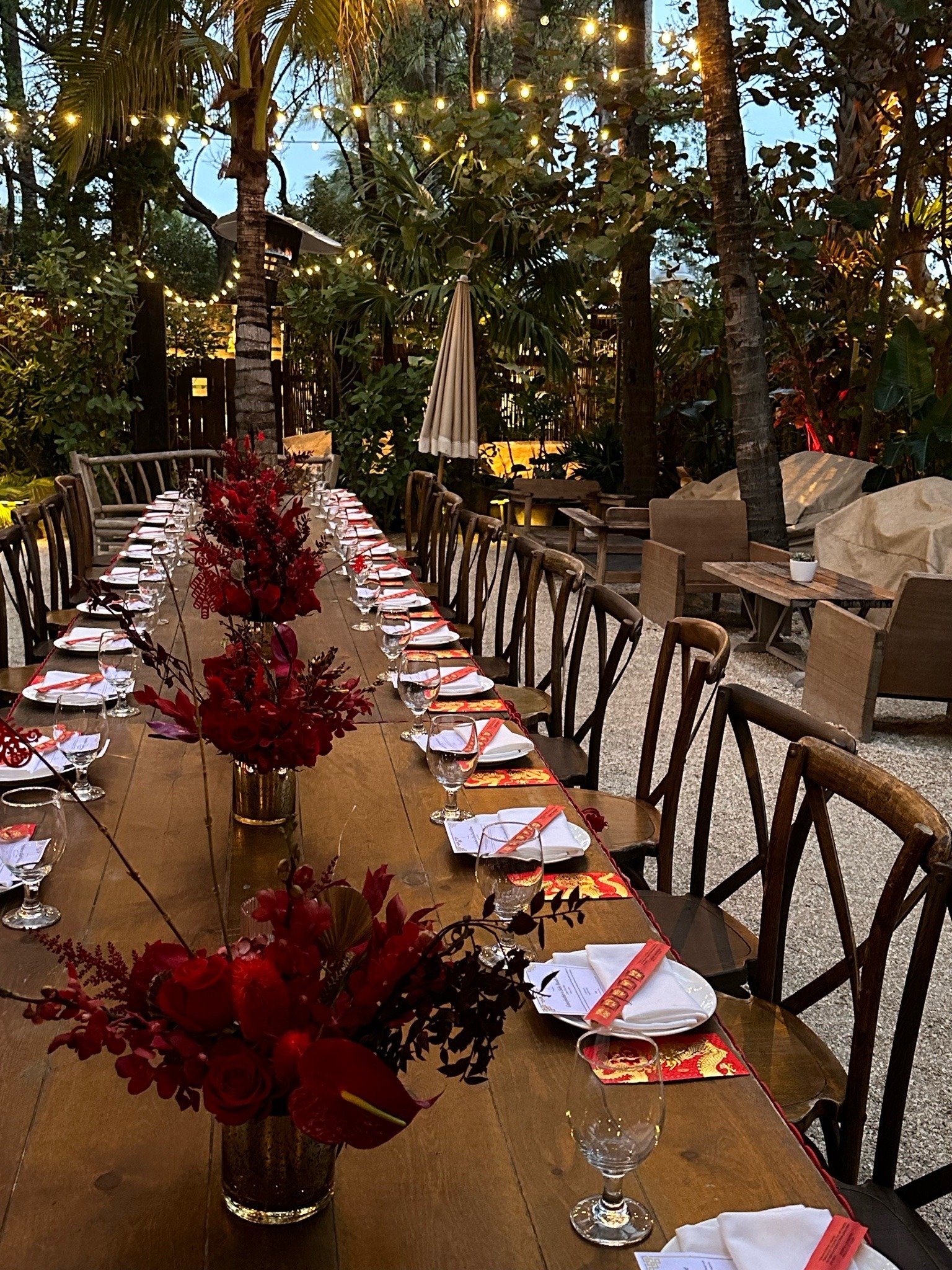 Long outdoor dining table set for a meal with floral centerpieces, wine glasses, napkins, and menus, surrounded by trees and string lights in an outdoor garden setting in the evening.