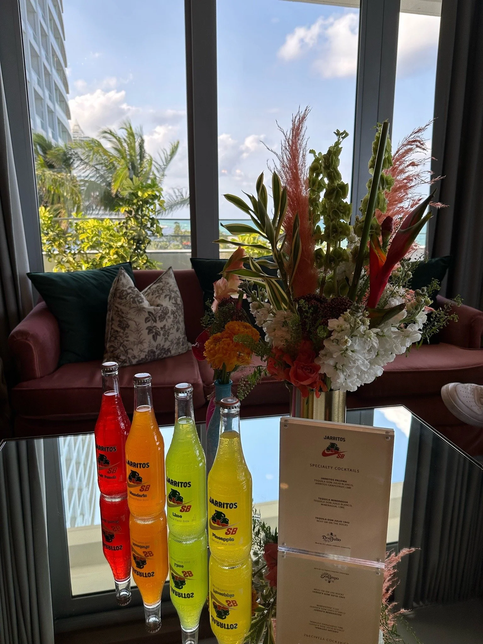 A glass-top table with bright multicolored bottles of Jarritos soda, a floral arrangement in a gold vase, and a menu card, with a settee and cushions in the background. Large windows reveal trees and a partly cloudy sky outside.