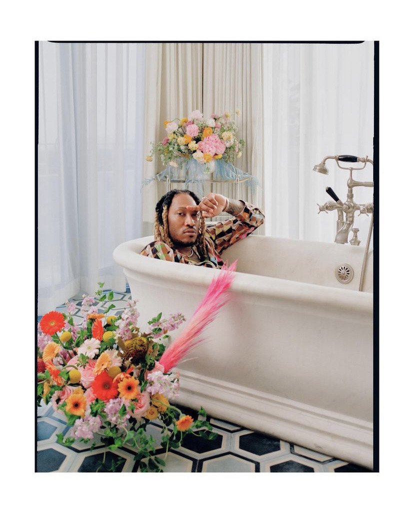 A man with long dreadlocks reclining in a vintage bathtub surrounded by colorful flowers, with a bouquet on a stand behind him and pouring pink pampas grass on the floor in front of him.