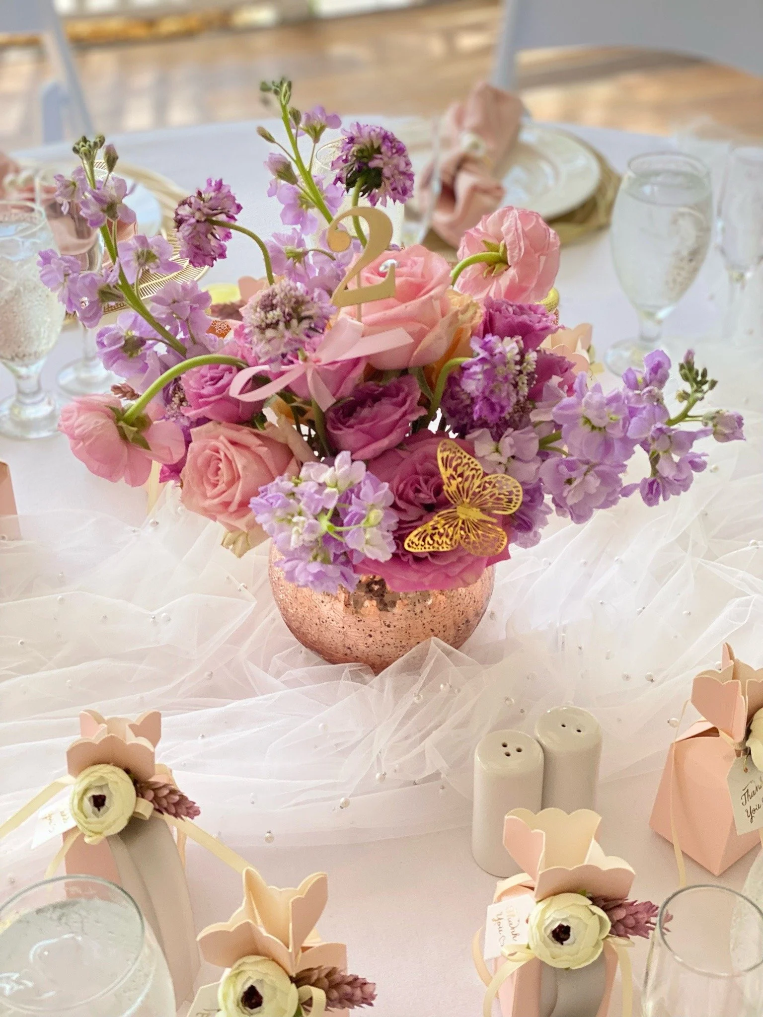 A pink and purple floral centerpiece with roses, delphiniums, and a butterfly on a decorated table for a celebration.