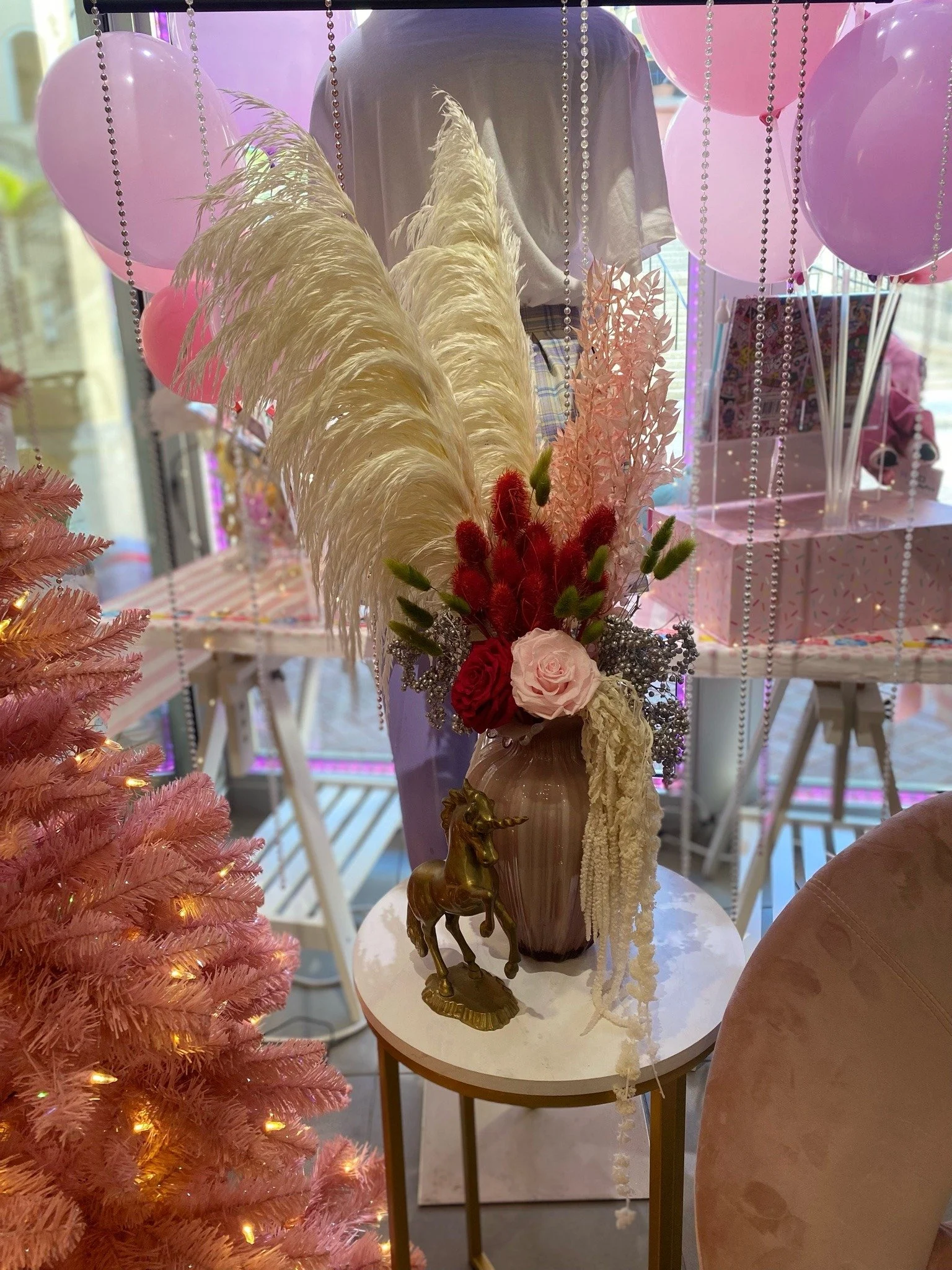 Decorative flower arrangement with pampas grass, roses, and other flowers in a vase, placed on a small white table next to a pink Christmas tree, with pink balloons and gift boxes in the background.