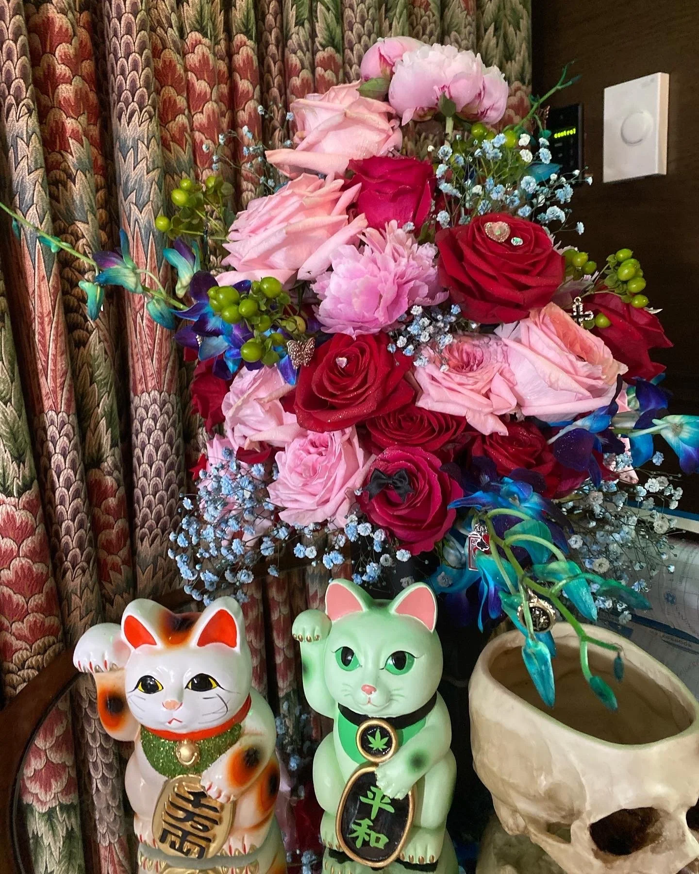 Colorful bouquet of pink, red, and purple flowers with two fortune cat figurines, one white with orange spots and the other green, in front of a patterned curtain and a decorative skull planter.