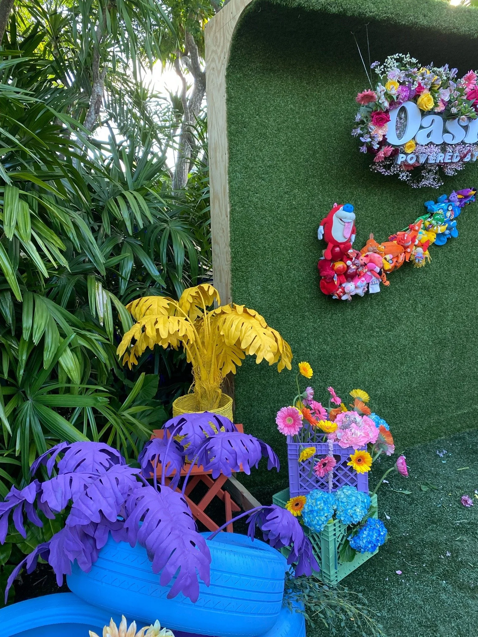 Colorful decorations with plush toys, flowers, and a sign that says "Oasis Powered" on a green artificial turf wall surrounded by lush green plants.