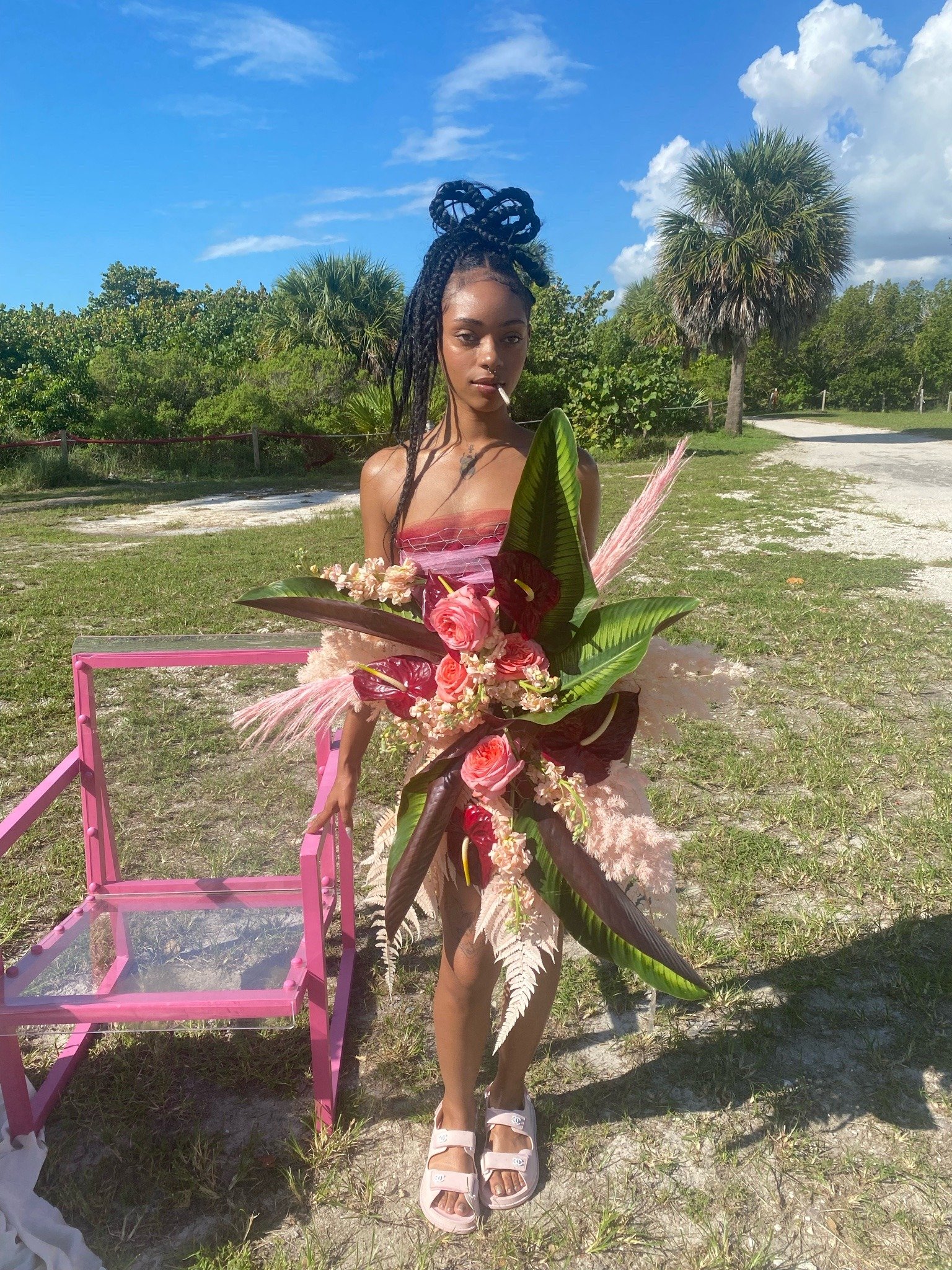 A young woman with braided hair styled in a high bun, standing outdoors on a sunny day with a background of palm trees and a blue sky. She is wearing a pink strapless dress and holding a large bouquet of pink and red flowers with green leaves, while standing next to a pink chair.