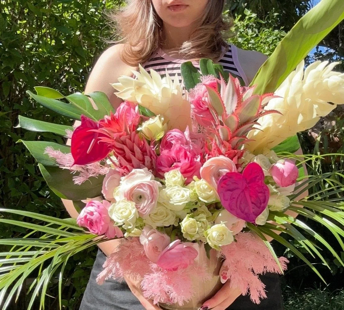 A woman holding a large colorful flower bouquet, featuring pink, white, and cream flowers with green leaves, outdoors in front of green foliage.