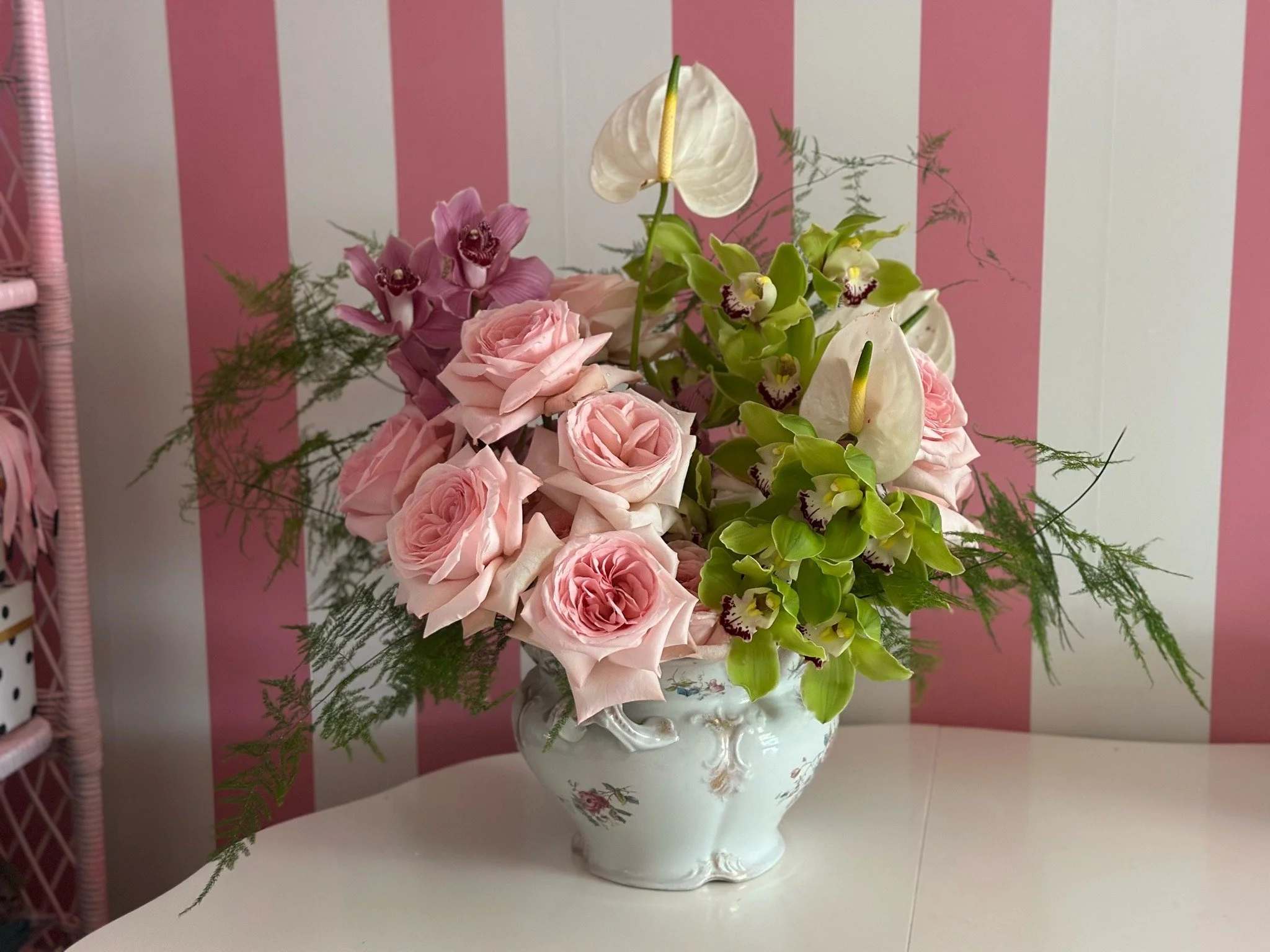 A floral arrangement in a white vintage-style vase featuring pink roses, white calla lilies, and green orchids, with fern-like greenery against a pink and white striped background.