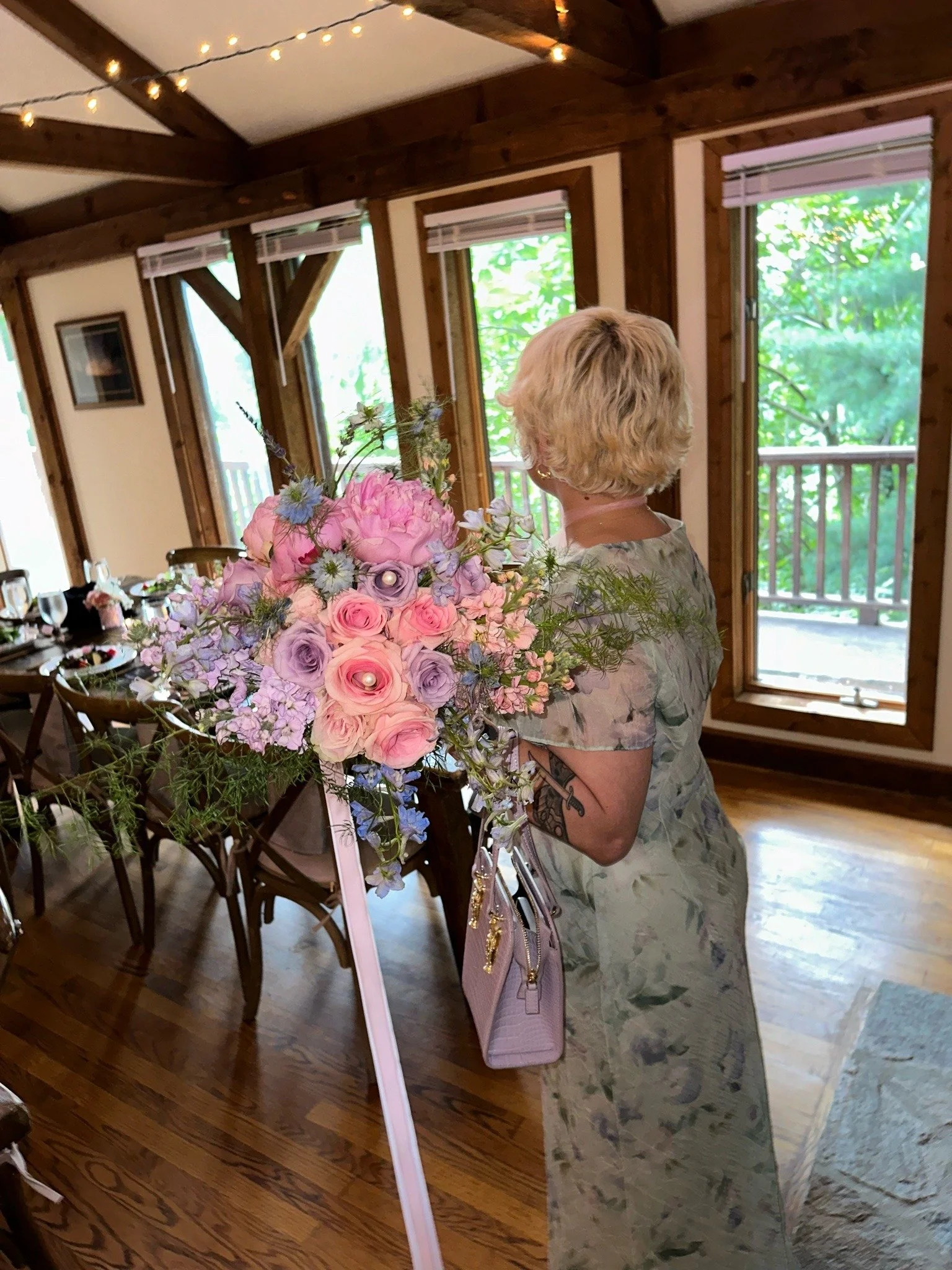 A woman with blonde hair and a floral dress holding a large bouquet of pink, purple, and white flowers in a wooden room with windows and a decorated table.