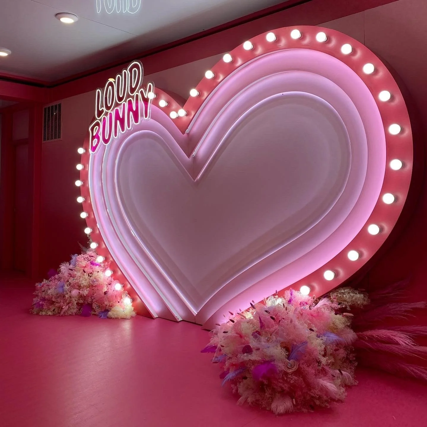 A large, illuminated pink heart-shaped display with the words 'Loud Bunny' in pink neon lights, decorated with fairy lights and floral arrangements at the base.