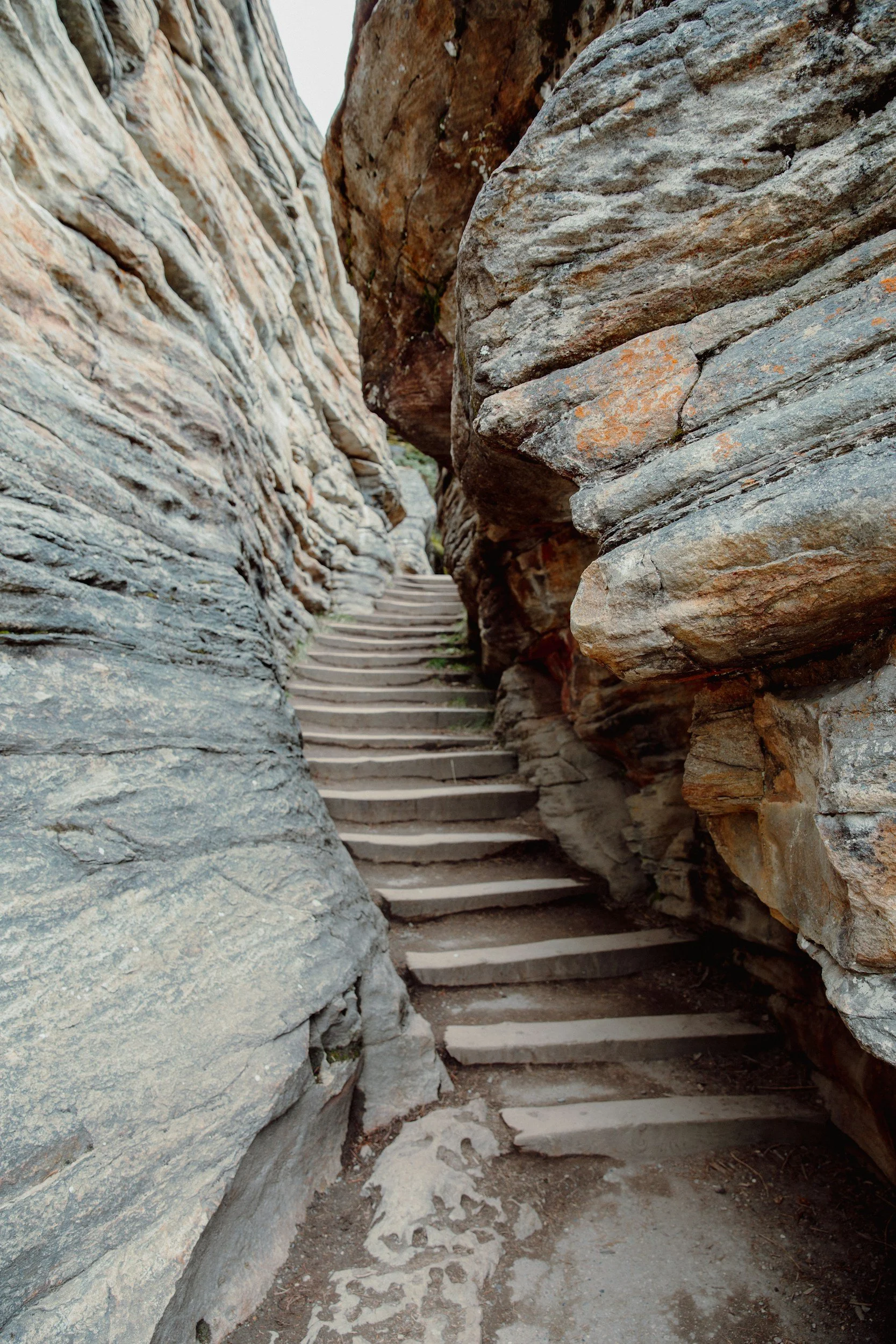 Narrow stone staircase with wooden steps, flanked by large, weathered rocks on both sides, leading upward into a rocky pathway.