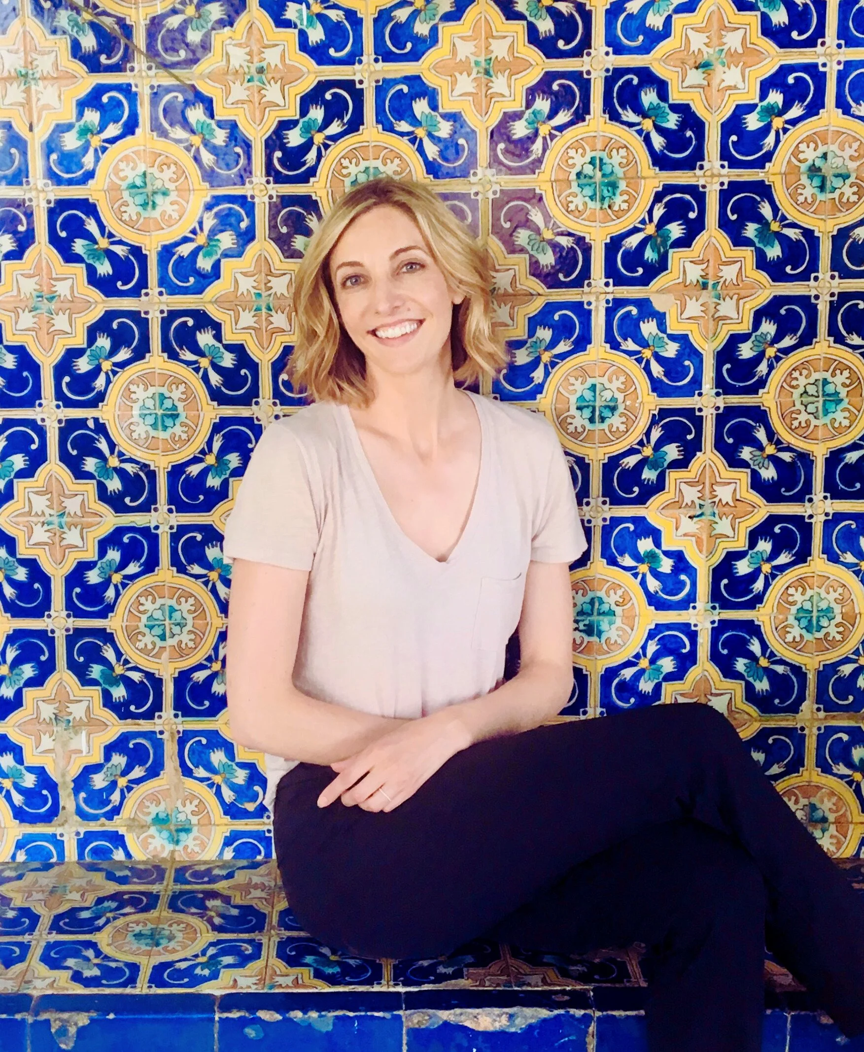 A woman with shoulder-length blonde hair and a light beige top sitting in front of a decorative ceramic tile wall, smiling and winking.