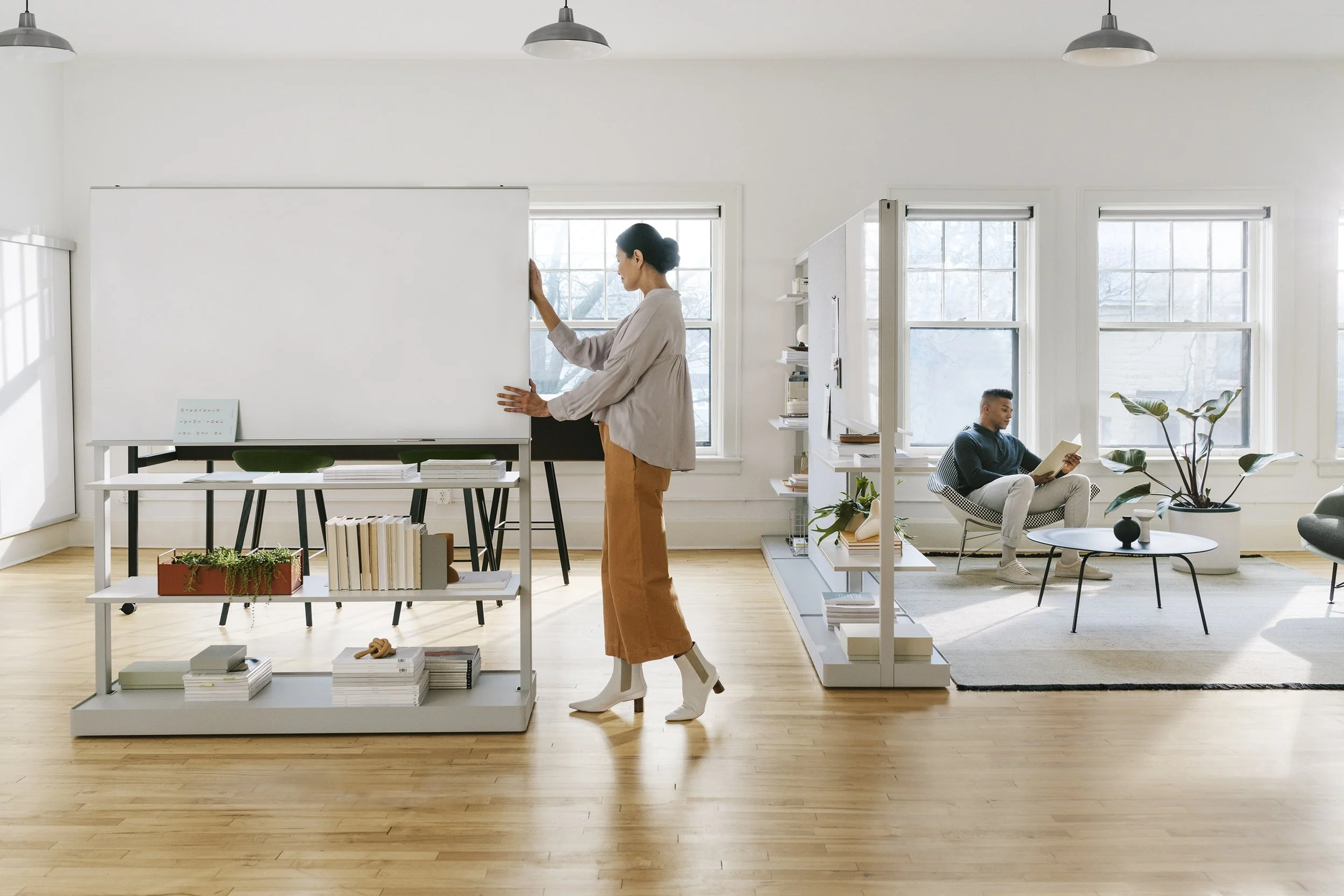 Woman standing near a whiteboard in a bright room with large windows. Man sitting on a chair reading a book in a seating area with a coffee table and potted plants.