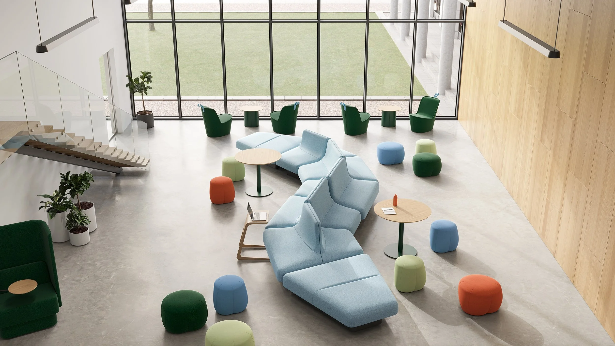 Modern, spacious lobby with light blue sectional sofa, various colorful ottomans, green armchairs, and small round tables. Large windows reveal a grassy outdoor area, and a staircase with glass railing is visible on the left. Potted plants decorate the space, and wooden wall panels add warmth.