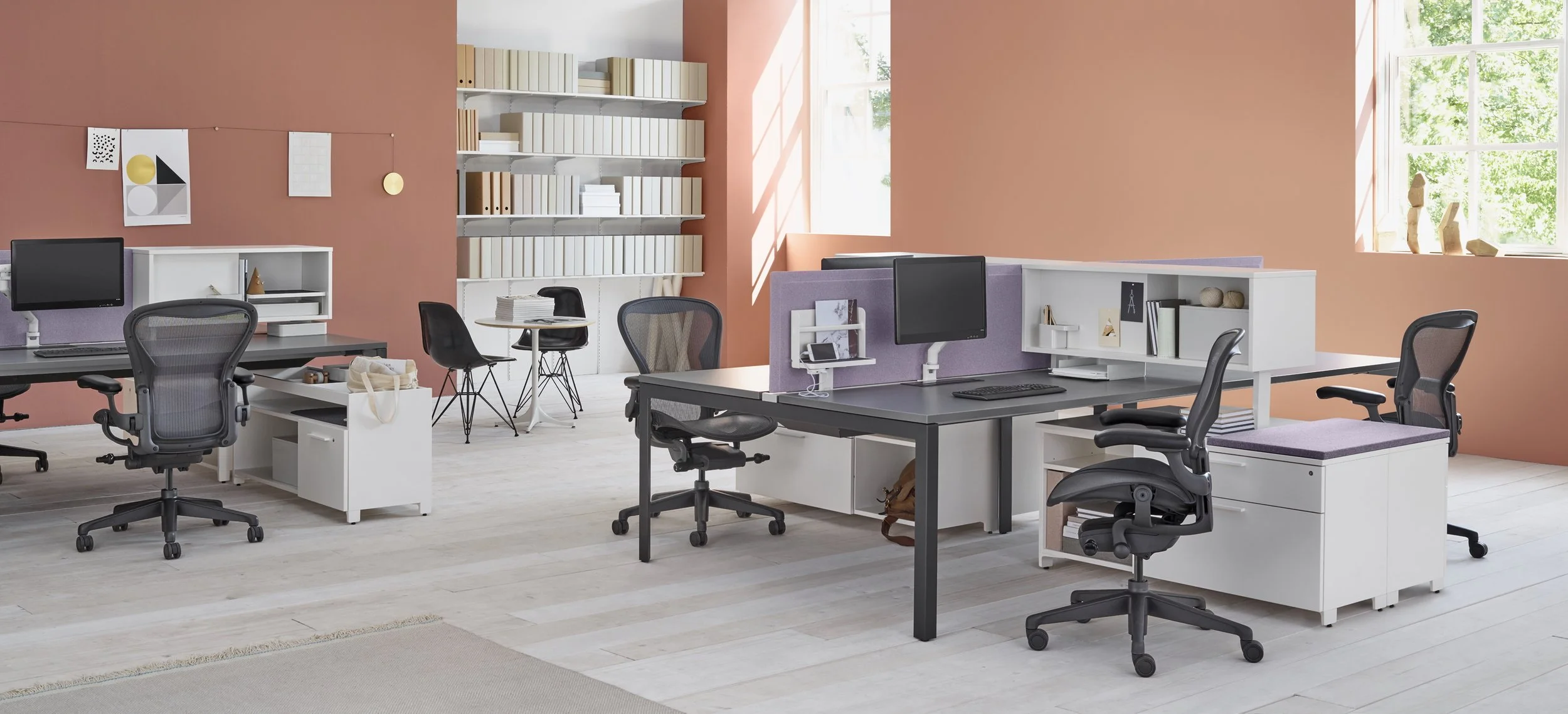 An office space with multiple desks, chairs, and computer monitors, featuring pink and purple accents and large windows letting in natural light.