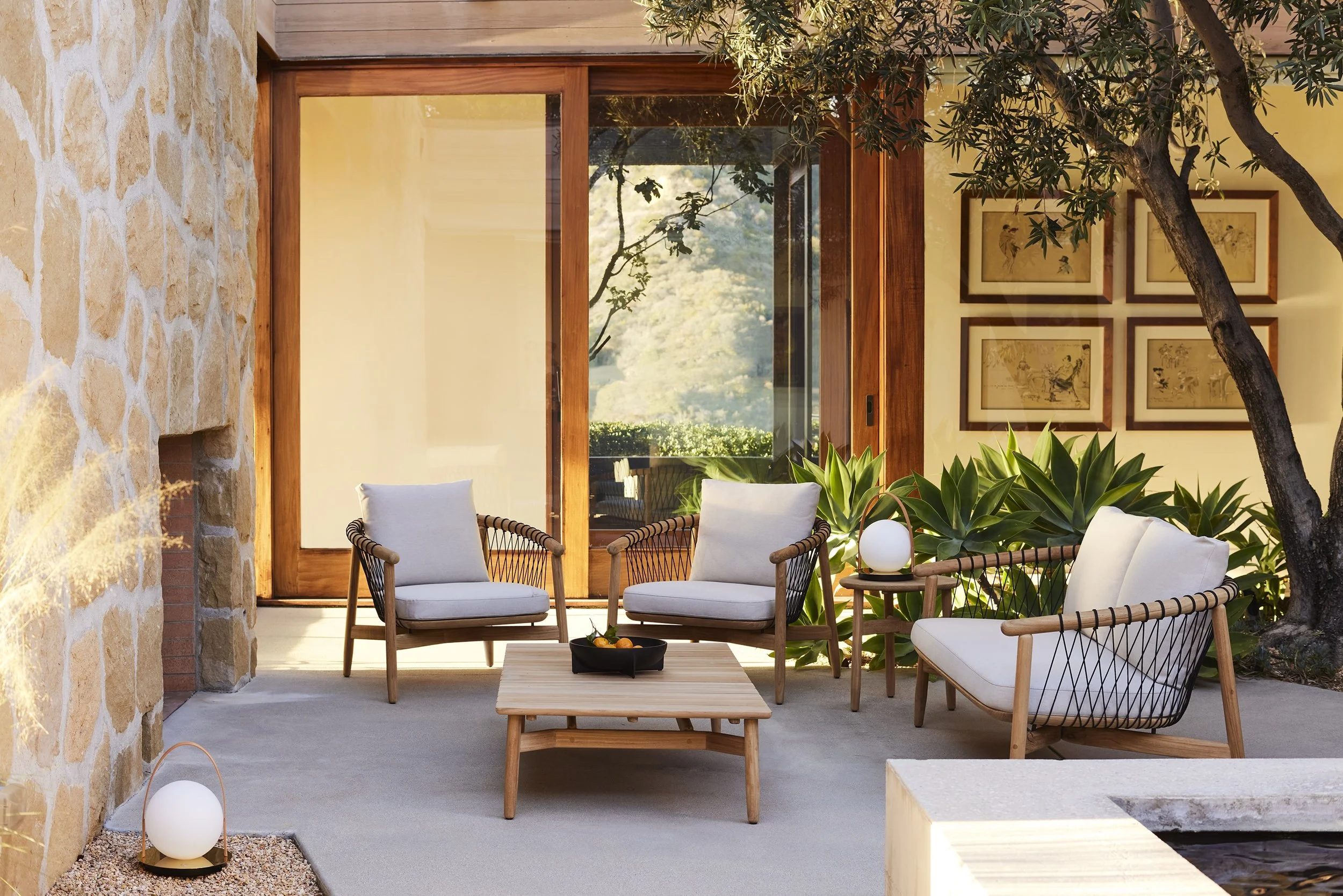 Outdoor patio with wooden furniture, two armchairs with white cushions, a wooden coffee table, and a side table with a round lamp. Large green plants and trees are visible, with a glass door and framed art on the interior wall.