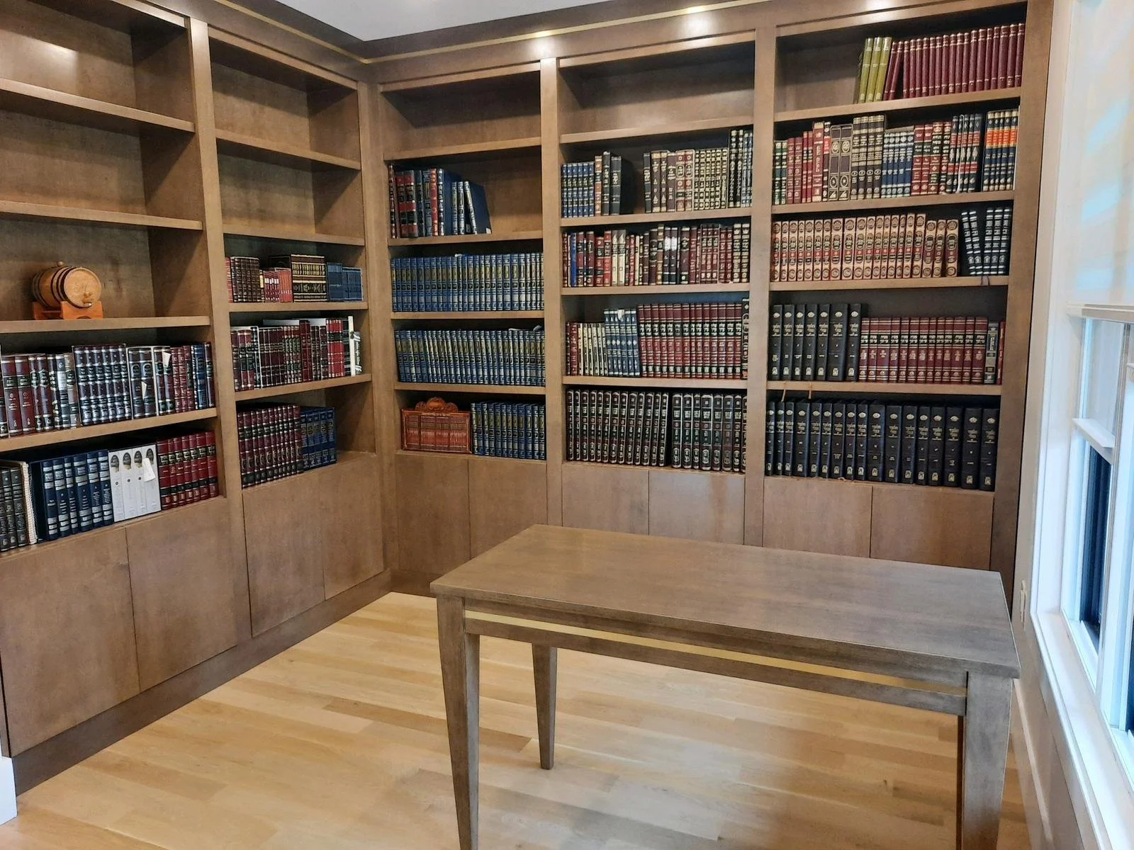 A wooden bookshelf filled with legal books, a wooden table, and a window with white blinds in a room with hardwood floors.